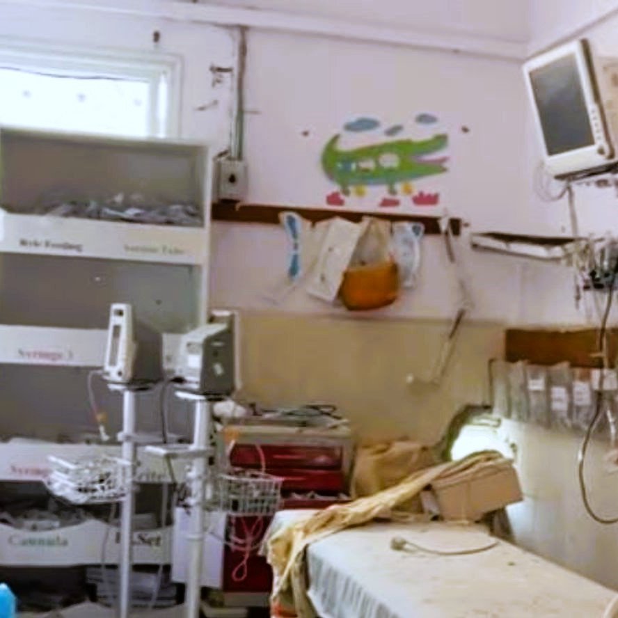 L'image montre une chambre d'hôpital qui a l'air un peu désordonnée et ancienne. On y voit un lit médical avec des draps, une étagère avec des boîtes, ainsi que du matériel médical. Un petit téléviseur est fixé au mur, et des illustrations colorées sont visibles sur les murs, ce qui ajoute une touche enfantine à l'environnement. La pièce semble nécessiter un peu de nettoyage et de modernisation.