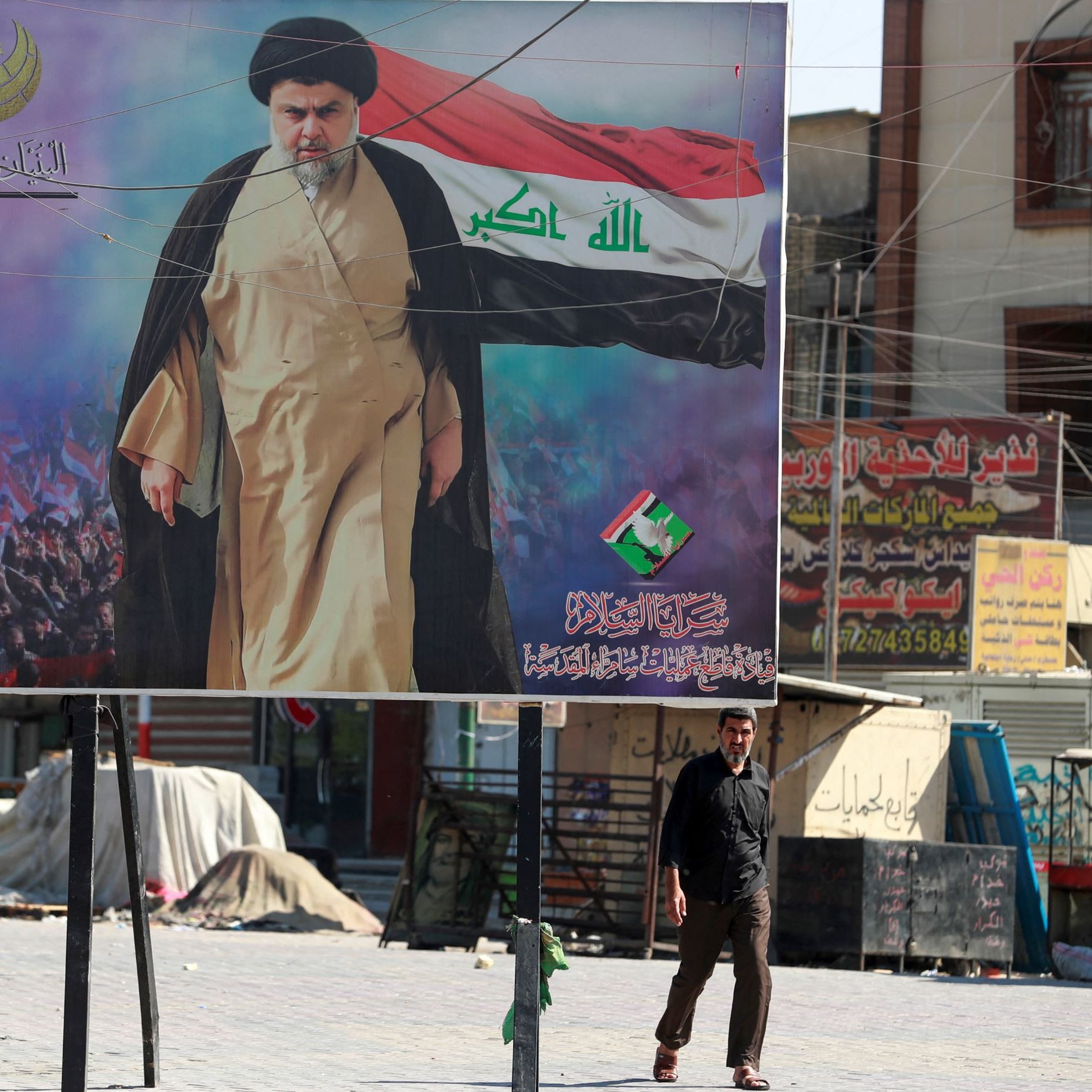 در این تصویر یک بیلبورد بزرگ نشان داده شده است که یک مرد با لباس روحانی و مسئولیت‌های سیاسی را در حال حرکت به سمت دوربین به تصویر می‌کشد. او دست خود را بلند کرده و یک پرچم در پس‌زمینه وجود دارد که نشان‌دهنده نمادها و پیام‌های خاصی است. در پس‌زمینه، جمعیتی از مردم در حال تماشا و حمایت هستند. فضای عمومی تصویر نشان‌دهنده یک منطقه شهری است که شاید تحت تأثیر مسائل اجتماعی یا سیاسی باشد. 