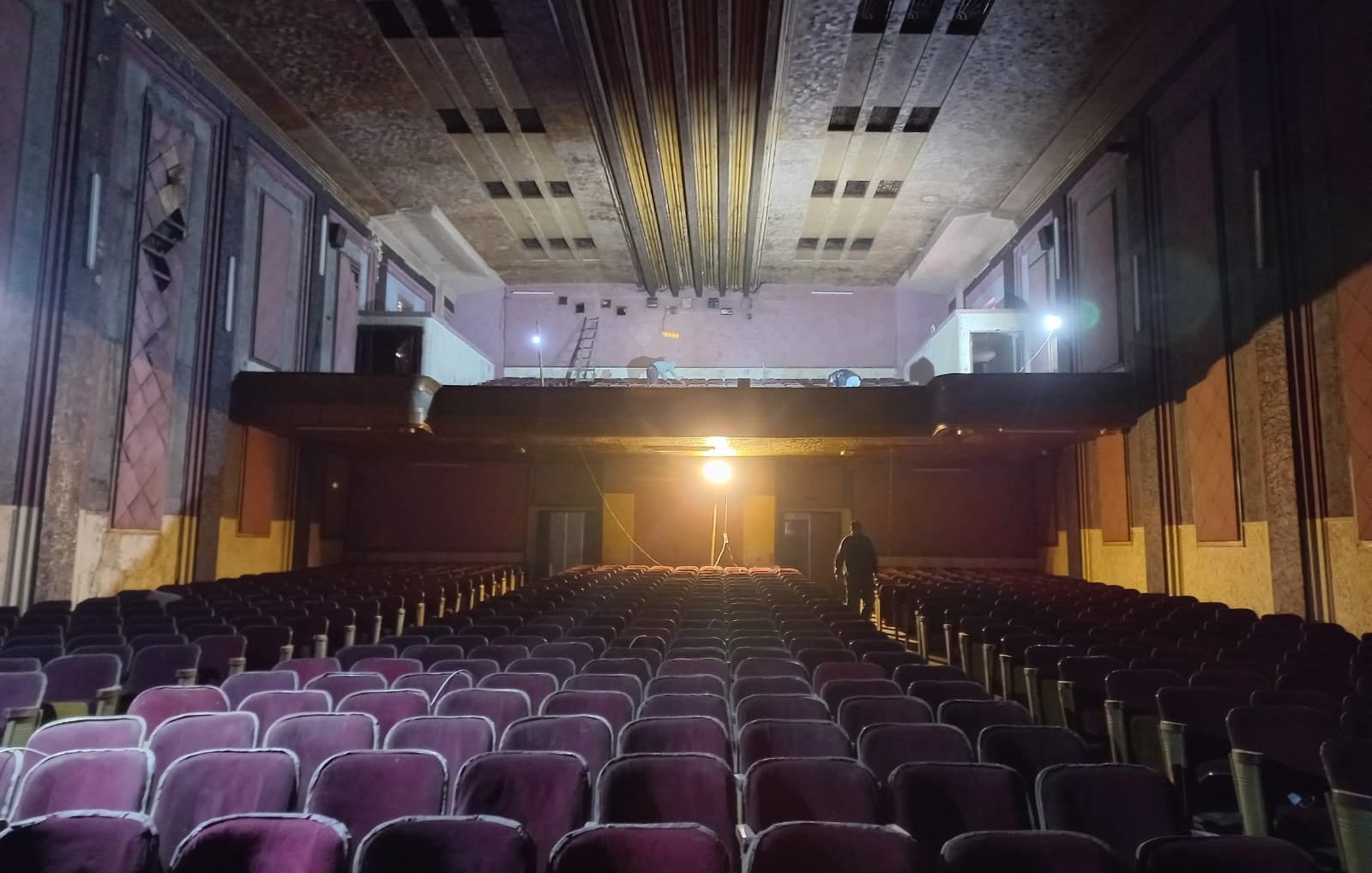L'image montre l'intérieur d'un théâtre ancien. On peut voir des rangées de sièges en velours, qui semblent un peu usés, dirigées vers une scène surélevée. La scène est éclairée par une lumière vive, tandis que le reste de la salle est dans une ambiance plus sombre. On aperçoit également des détails architecturaux au plafond et sur les murs, qui évoquent un style vintage. Un homme se tient debout au fond, ajoutant une touche humaine à cet espace, qui dégage une atmosphère à la fois nostalgique et mystérieuse.