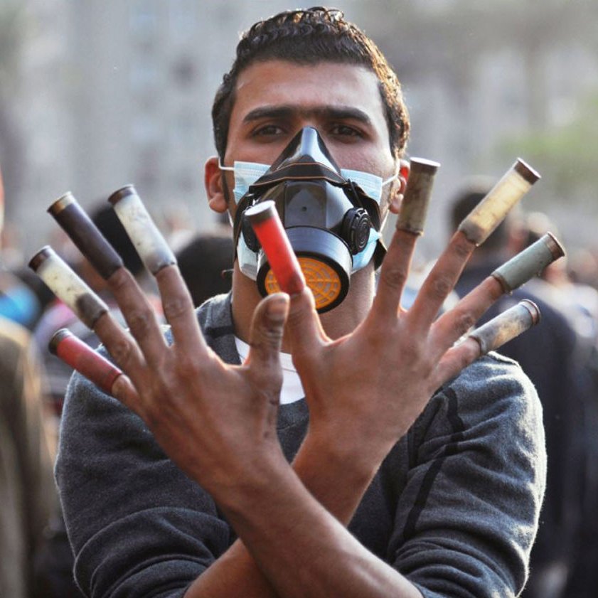 L'image montre un homme portant un masque de protection, levant les mains avec plusieurs cartouches de gaz lacrymogène visibles. Son expression semble déterminée, et l'arrière-plan indique qu'il est présent dans une foule, probablement lors d'une manifestation. L'atmosphère de la scène semble être celle d'un événement intense et chargé d'émotion.