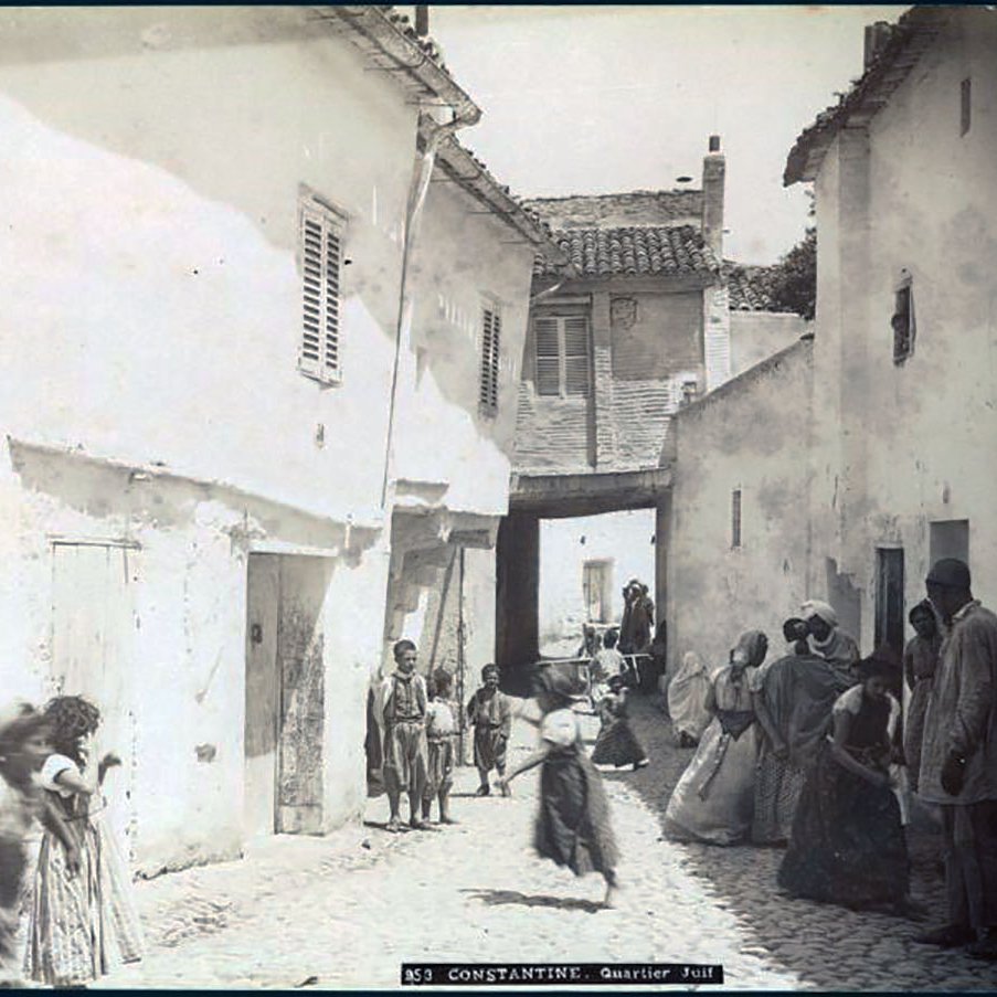 L'image représente une scène de rue dans un quartier de Constantine, en Algérie. On y voit des maisons blanches avec des volets, et une ruelle pavée qui semble étroite. Plusieurs personnes, y compris des enfants, sont visibles, certaines marchant et d'autres se tenant debout. Les vêtements des habitants semblent traditionnels, reflet d'une époque passée. L'atmosphère générale évoque une vie quotidienne animée dans un contexte historique.