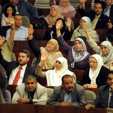 L'image montre un rassemblement dans un espace législatif, probablement au sein d'une assemblée ou d'un parlement. De nombreuses personnes sont présentes, avec une majorité levant la main, ce qui indique probablement un vote ou une approbation. Les participants semblent porter des tenues formelles, et certaines femmes portent le hijab. On observe une diversité de participants, tant en termes de genre que d'âge. L'environnement évoque une atmosphère de discussion et de prise de décision collective.