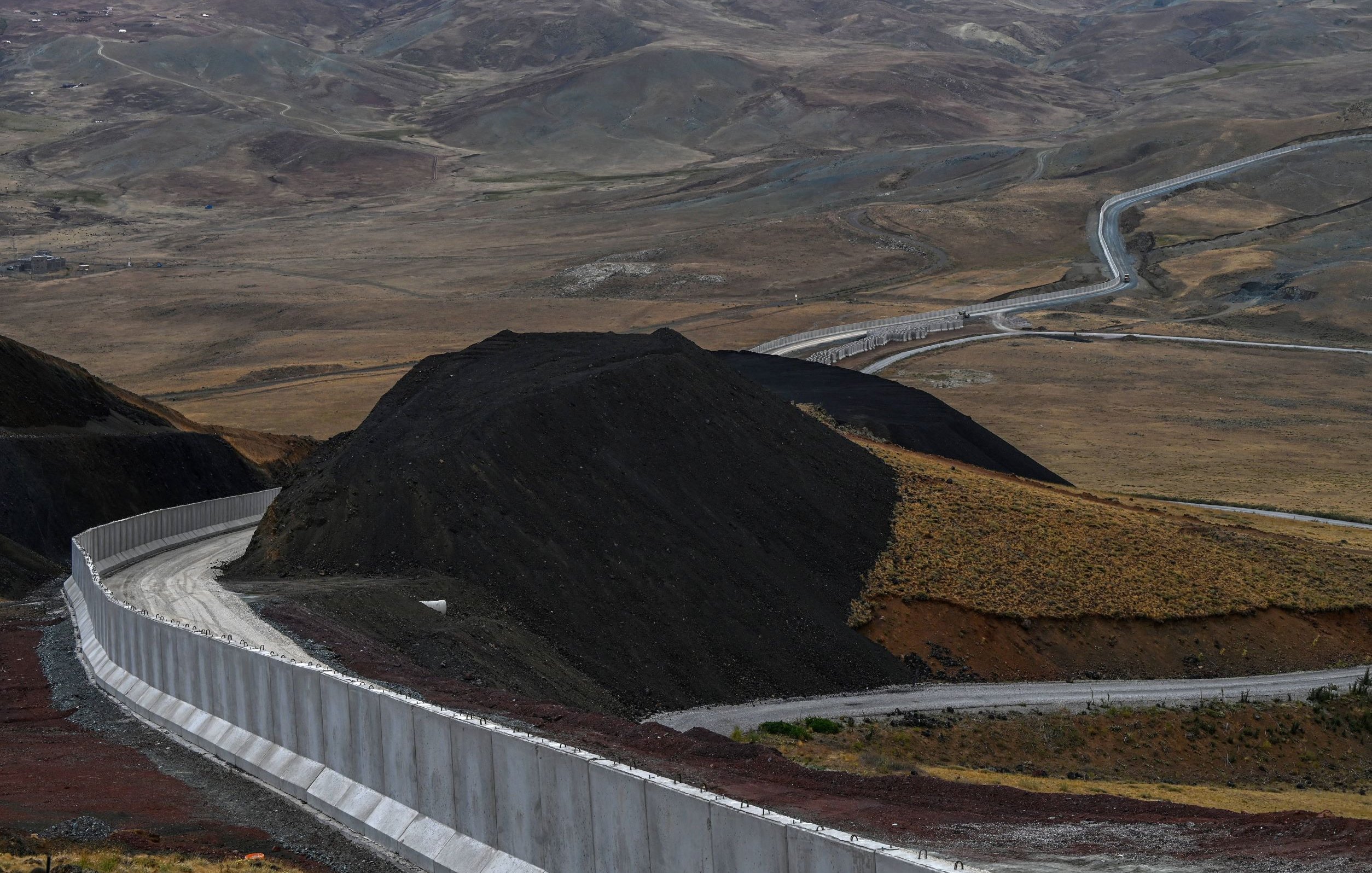 L'immagine mostra un paesaggio montano caratterizzato da una strada che si snoda attraverso un terreno collinare. Sullo sfondo, si possono vedere colline e valli, mentre in primo piano c'è una parete di cemento che segue il contorno della strada. Il terreno appare secco e brullo, con tonalità di marrone e verde. La scena comunica un senso di isolamento e vastità, tipico di un ambiente montano.