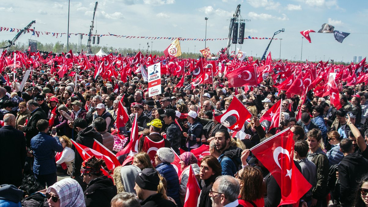 L'image montre une grande foule rassemblée en plein air, probablement lors d'un événement ou d'une manifestation en Turquie. De nombreuses personnes tiennent des drapeaux turcs, qui sont rouges avec un croissant blanc et une étoile. On peut voir des banderoles et des panneaux, et l'ambiance semble dynamique et engagée, avec un ciel bleu parsemé de nuages.