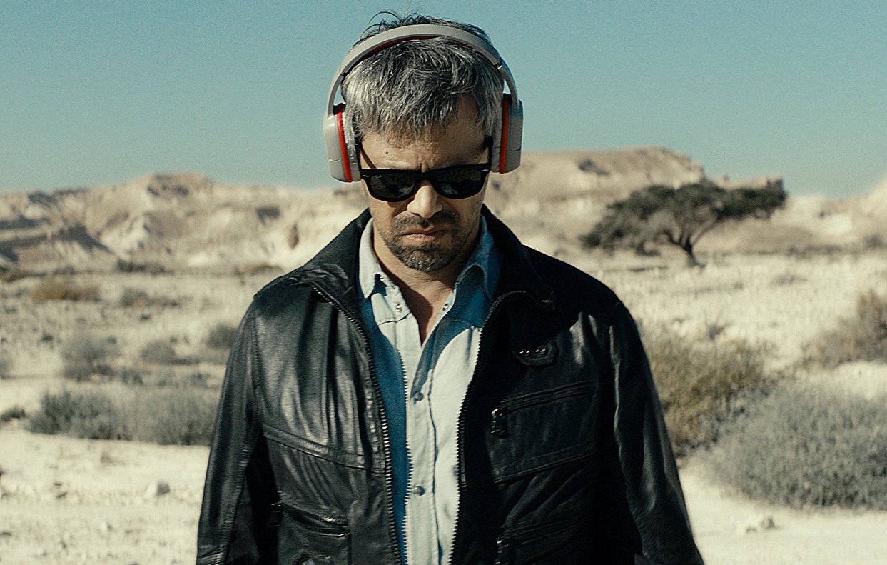 L'image montre un homme dans un paysage désertique. Il porte des écouteurs rouges et des lunettes de soleil, ce qui lui donne un air détaché. Il est vêtu d'une veste en cuir noire et semble perdu dans ses pensées, les yeux baissés. Le décor environnant est aride, avec des collines au loin et une atmosphère de solitude.
