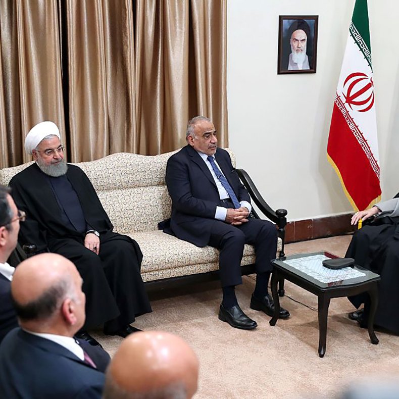 L'image montre une réunion formelle dans un bureau. Plusieurs personnalités sont assises, avec des expressions sérieuses, et se regardent. Au fond, on peut voir un drapeau iranien et un portrait accroché au mur. Les personnes sont vêtues de costumes professionnels, créant une ambiance de discussion diplomatique.