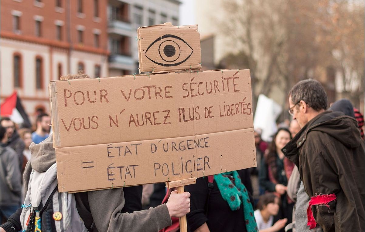 L'image représente une manifestation où une personne tient une pancarte en carton. Sur cette pancarte, on peut lire un message dénonçant la restriction des libertés au nom de la sécurité. Le symbole d'un œil est visible en haut de la pancarte, renforçant le thème de la surveillance. Les participants de la manifestation semblent mobilisés, et il y a une ambiance de protestation dans l'air.