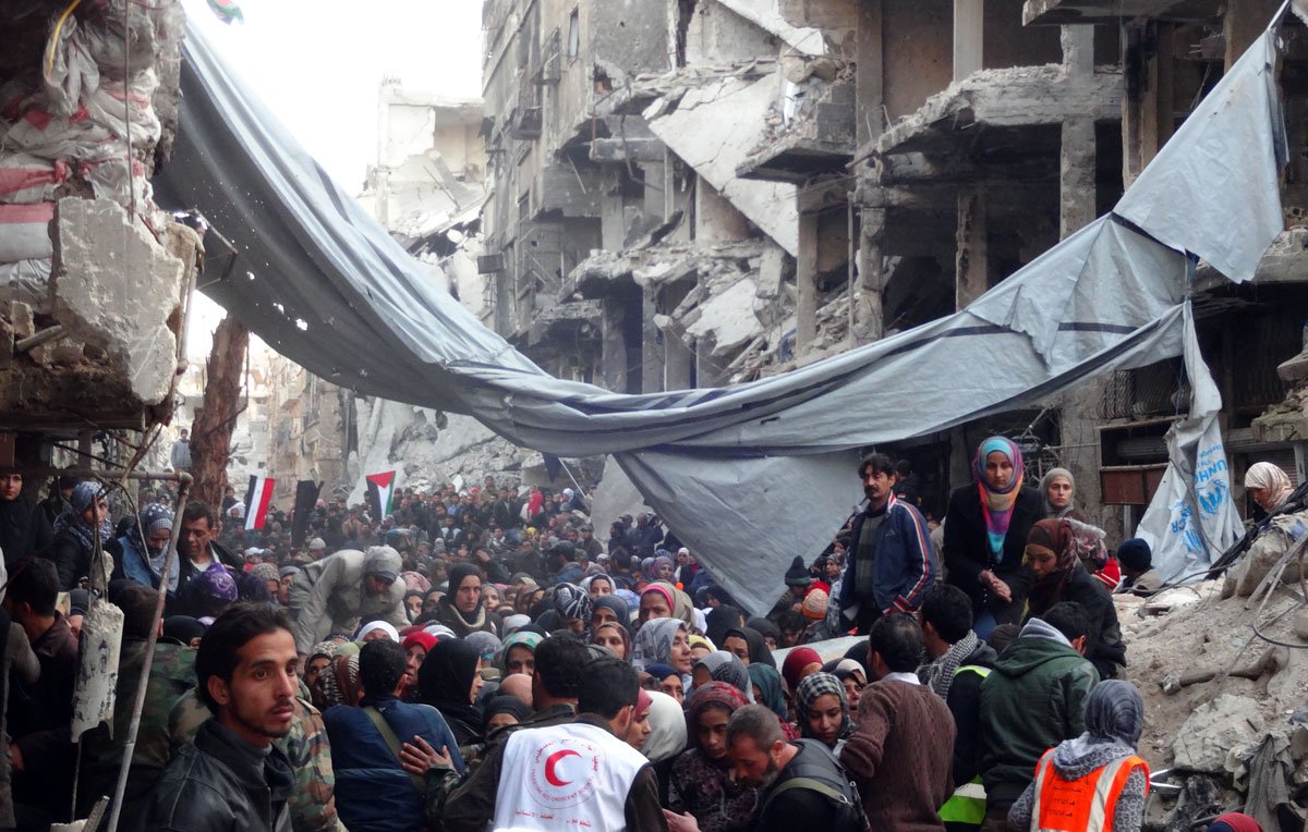 L'image montre une scène de destruction massive dans une zone urbaine, probablement après un conflit ou une catastrophe. On peut voir des bâtiments gravement endommagés, avec des murs effondrés et des débris partout. Une foule de personnes, dont certaines semblent déplacées, se rassemble dans la rue. Il y a des tentes et des bâches qui servent d'abris temporaires. Des membres d'organisations humanitaires, identifiables par leurs vêtements distinctifs, sont présents pour aider. L'ambiance générale est celle de la détresse et de la recherche d'aide dans une situation de crise.