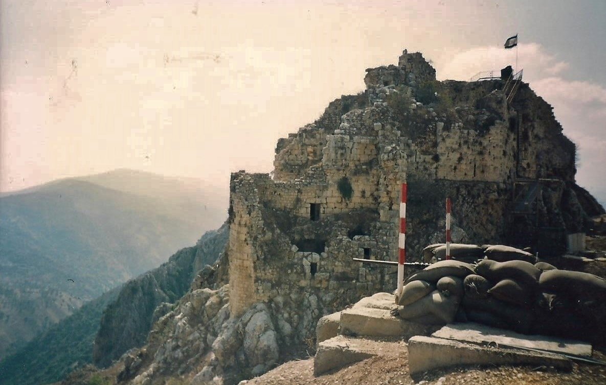 L'image montre les ruines d'une vieille forteresse perchée sur une colline, avec des murs en pierre partiellement érodés. Des drapeaux flottent au sommet de la structure. En premier plan, on peut voir des sacs de terre disposés en protection, suggérant une utilisation militaire. La vaste vallée et les montagnes se dessinent à l'arrière-plan, créant une ambiance de paysages montagneux majestueux. L'ensemble évoque une atmosphère historique et stratégique.