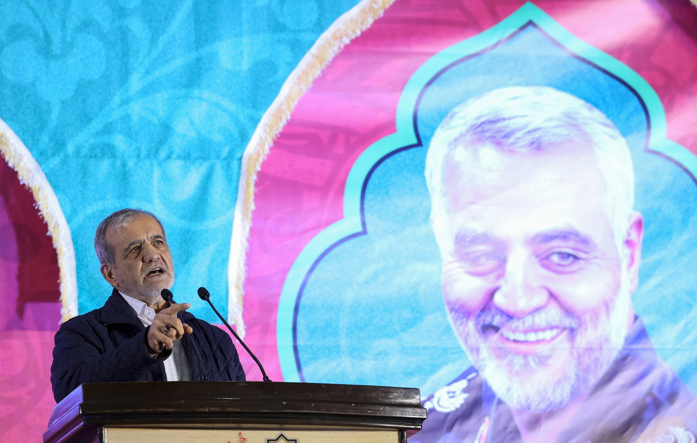 در این تصویر، شخصی در حال سخنرانی در یک مراسم دیده می‌شود. او پشت میکروفون قرار دارد و به طرف جمعیت صحبت می‌کند. در پس‌زمینه، تصویری بزرگ از فرد دیگری که لباس نظامی به تن دارد، به‌وضوح قابل مشاهده است. طراحی پس‌زمینه رنگی و با الگوهای خاص است که به مراسم زیبایی می‌بخشد. فضای کلی مراسم جدی و متعهد به هدف خاصی به نظر می‌رسد.