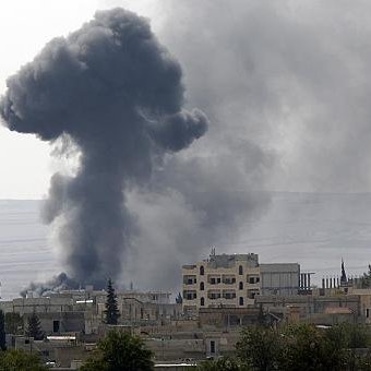 L'image montre un paysage urbain avec un nuage de fumée noir s'élevant dans le ciel, probablement à la suite d'une explosion ou d'un incendie. Les bâtiments aux alentours semblent endommagés, et la végétation est rare. Le fond présente des collines dégradées, ce qui suggère un environnement difficile. L'atmosphère générale évoque une situation de conflit ou de crise.