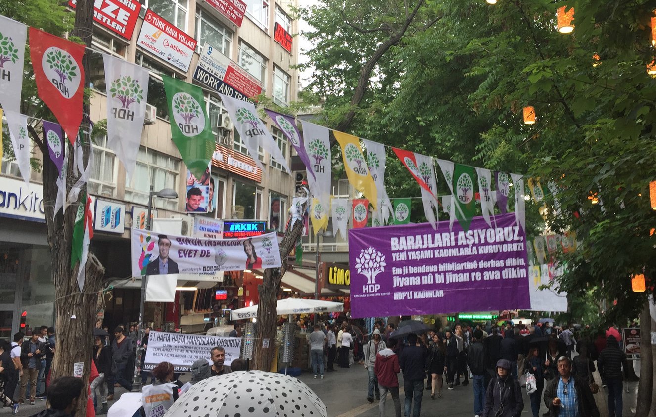 L'image montre une rue animée avec des banderoles accrochées entre les bâtiments. Ces banderoles semblent promouvoir un événement ou une cause, avec des slogans en caractères visibles. On peut également voir des groupes de personnes se rassembler au sol, suggérant une atmosphère de manifestation ou de réunion. Des arbres bordent la rue, ajoutant une touche de verdure à la scène. La journée semble nuageuse, ce qui pourrait indiquer un temps frais.