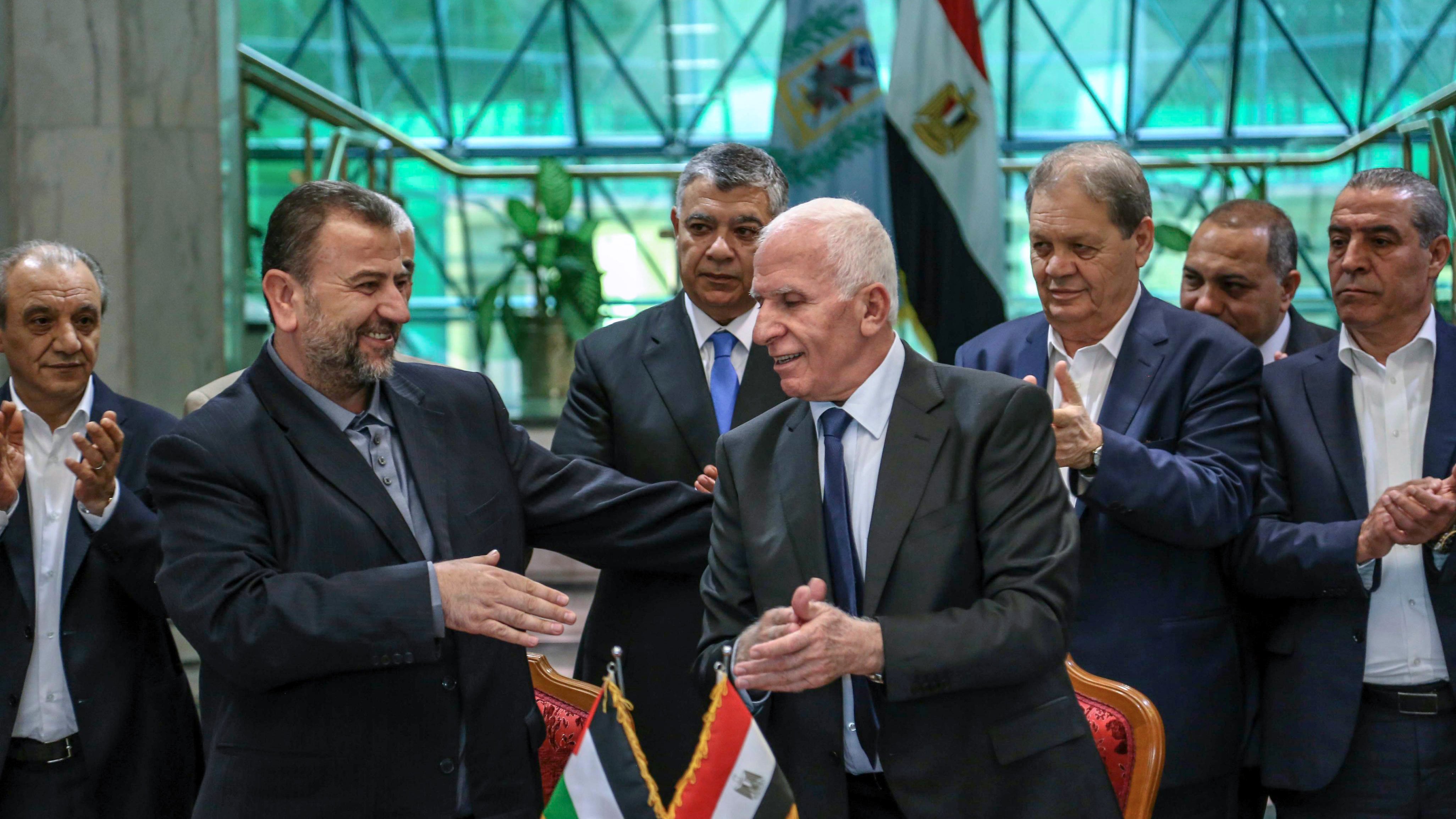 The image shows a formal gathering where two men are shaking hands, likely after a significant agreement or event. They are standing at a table with flags of Palestine and Egypt displayed in front of them. Behind them, a group of men appears to be applauding or showing support. The setting looks official, possibly within a government building, with a modern architectural backdrop. The atmosphere seems positive, indicating a moment of cooperation or partnership.
