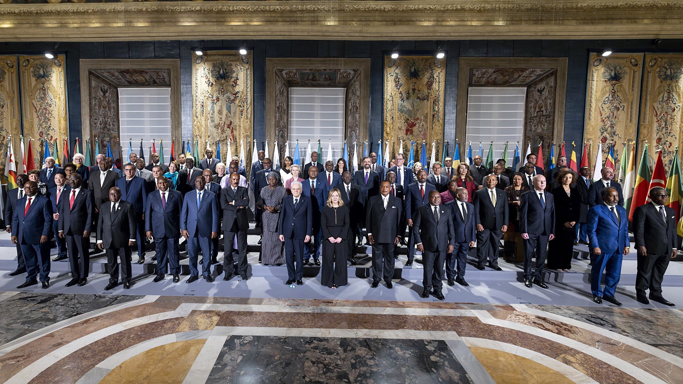 L'immagine mostra un gruppo di persone in un ambiente ufficiale, probabilmente durante un incontro o una conferenza internazionale. Si vedono diversi uomini e donne in abiti formali, che rappresentano vari paesi, dato che ci sono bandiere nazionali esposte sullo sfondo. L'atmosfera sembra seria e solenne, con tutti i partecipanti schierati in modo ordinato. Le decorazioni e l'architettura dell'ambiente circostante danno un tocco di grandezza all'evento.