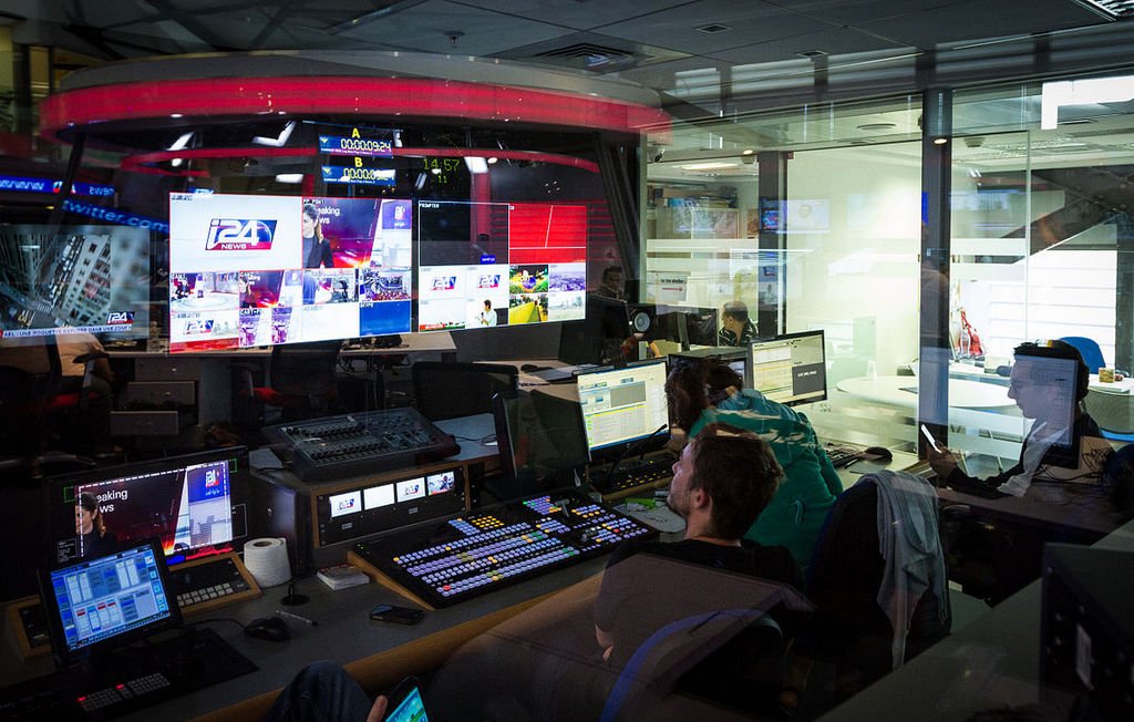 L'image montre une salle de contrôle de télévision ou de production, probablement un centre de diffusion. On y voit plusieurs écrans affichant des images en direct, des graphiques et des informations. Des personnes sont assises devant des consoles de contrôle, concentrées sur leur travail, et semblent superviser la diffusion. L'éclairage est tamisé, et on peut apercevoir une ambiance technologique avec des équipements modernes. Les murs en verre permettent d'apercevoir à l'extérieur, donnant une sensation d'ouverture.