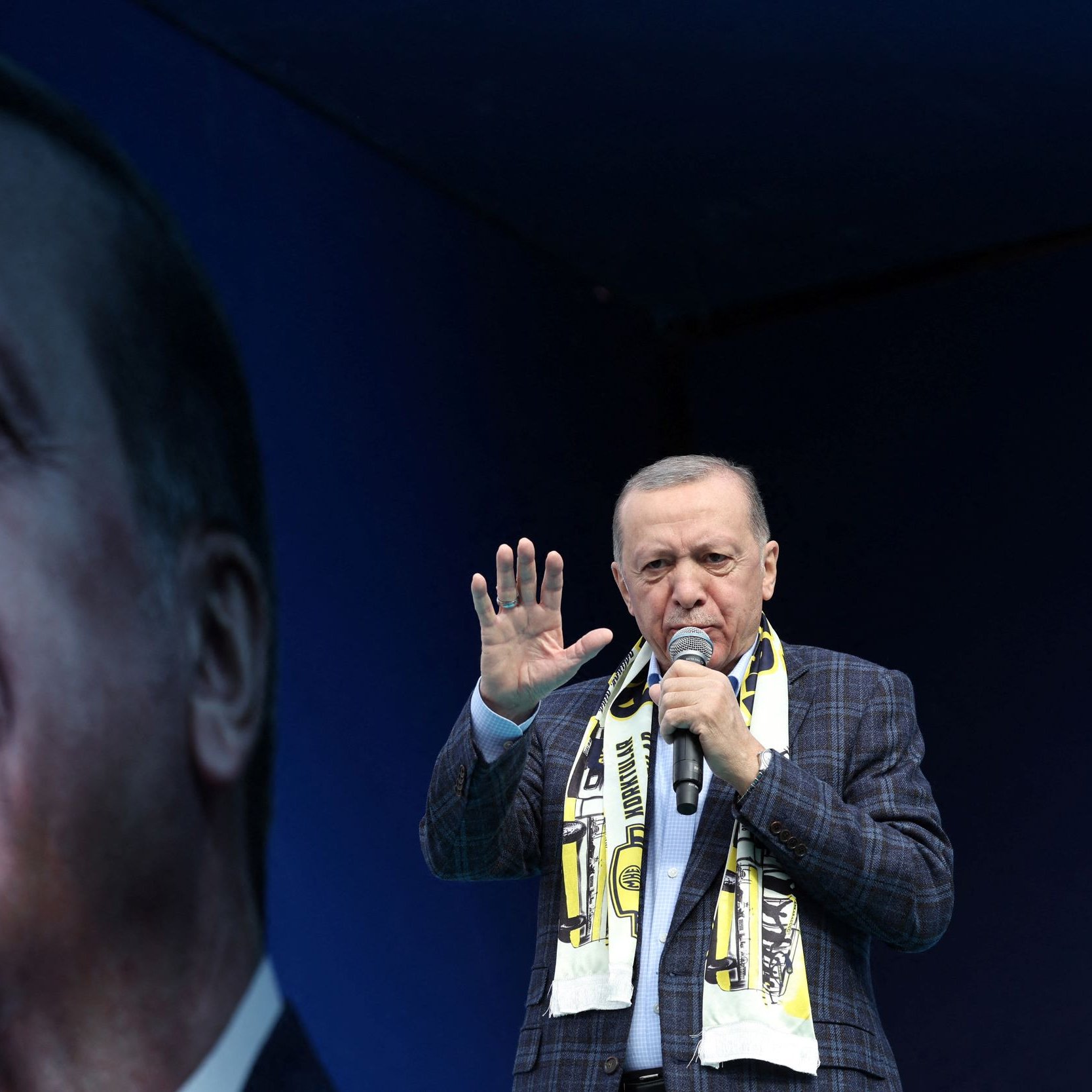 L'image montre un homme sur une scène, tenant un microphone et s'adressant à une foule. Il porte une écharpe aux couleurs d'une équipe de football et semble faire un discours passionné. En arrière-plan, on peut voir un grand portrait de lui, qui renforce sa présence sur scène. La scène est ornée d'un fond bleu, mettant en avant son image.