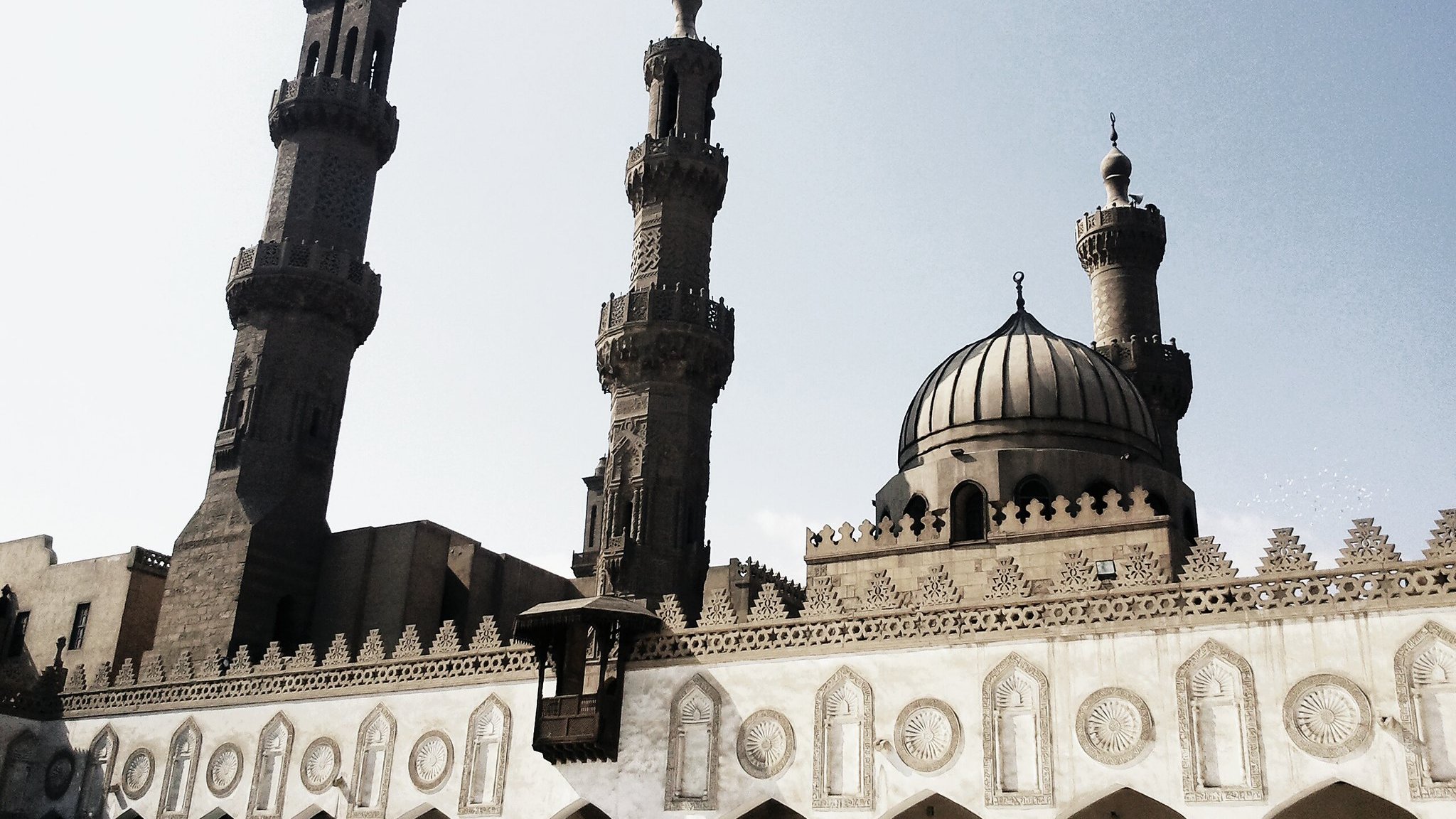 L'image montre une mosquée majestueuse avec deux minarets qui s'élèvent haut dans le ciel. La structure présente une belle architecture islamique, caractérisée par des arches décoratives et des motifs ornementaux sur les murs. Le dôme au centre ajoute à la grandeur du bâtiment, tandis que le ciel clair en arrière-plan donne une ambiance lumineuse à la scène. Les détails sculptés et la symétrie des éléments architecturaux témoignent d'un riche héritage culturel.