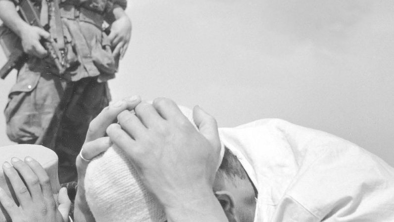 L'image montre un soldat en uniforme tenant une arme, se tenant en arrière-plan, tandis que deux personnes au premier plan sont inclinées vers le sol. L'une d'elles semble tenir sa tête dans ses mains, exprimant une grande détresse ou un sentiment de soumission. L'ambiance générale semble chargée d'émotion et de tension, suggérant une situation de conflit ou de crise. Les éléments monochromes de la photographie renforcent l'intensité de la scène.