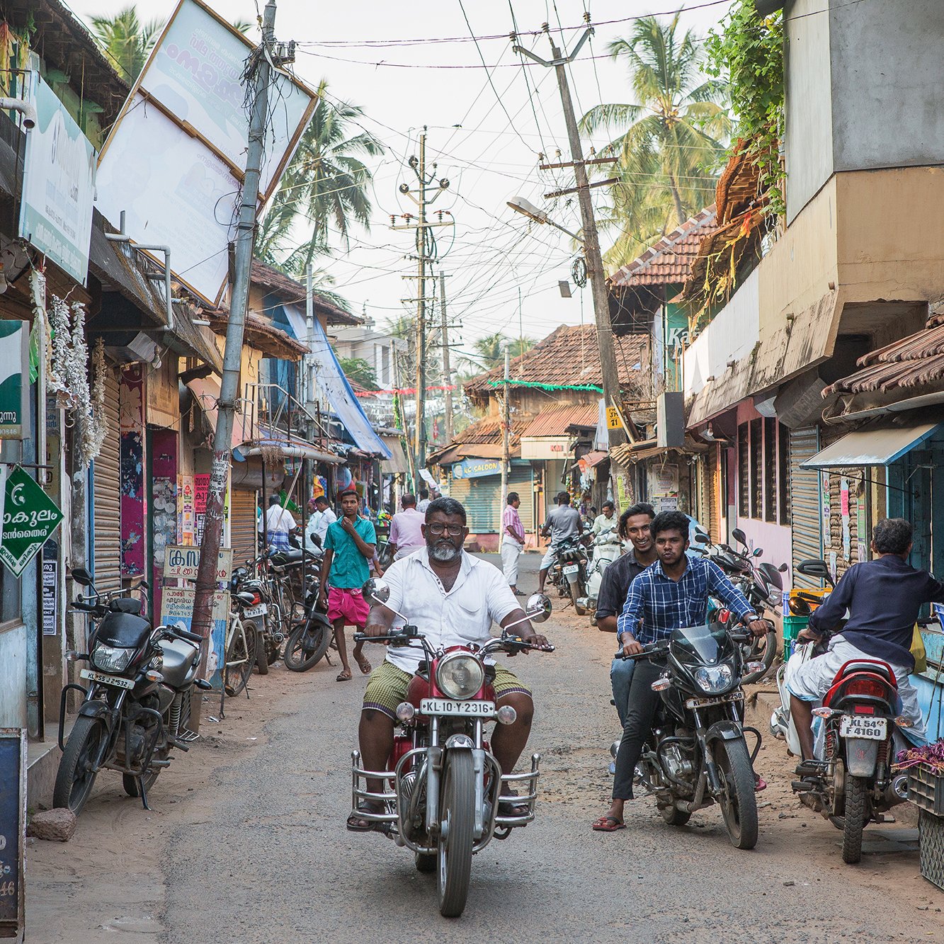 L'image montre une rue animée d'un village ou d'une petite ville. On y voit des bâtiments aux façades colorées et typiques de l'architecture locale, avec des toits en tuiles. Plusieurs personnes sont présentes, certaines se déplaçant à pied, d'autres sur des motos. Des échoppes bordent la rue, vendant probablement des fruits et légumes. L'atmosphère semble vivante et animée, avec des fils électriques et des palmiers visibles en arrière-plan.