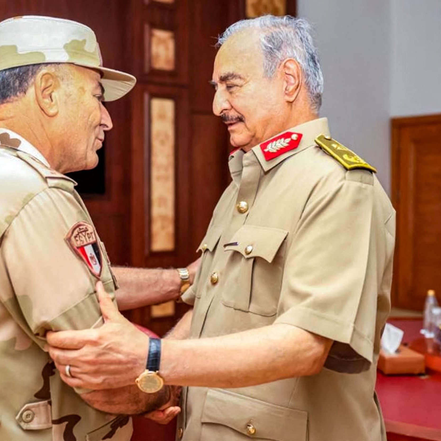 در این تصویر، دو شخص نظامی در حال گفتگو و تبادل نظر هستند. یکی از آن‌ها Uniform نظامی با رنگ‌های بیابانی و شَمشیر نشان‌دار پوشیده است و دیگری یونیفرم نظامی با رنگ‌های مشابه دارد. فضای محیط رسمی و دارای دکور چوبی است. به نظر می‌رسد که این دو نفر در یک جلسه یا دیدار رسمی حضور دارند و بین آن‌ها احترامی دوستانه و صمیمی برقرار است.