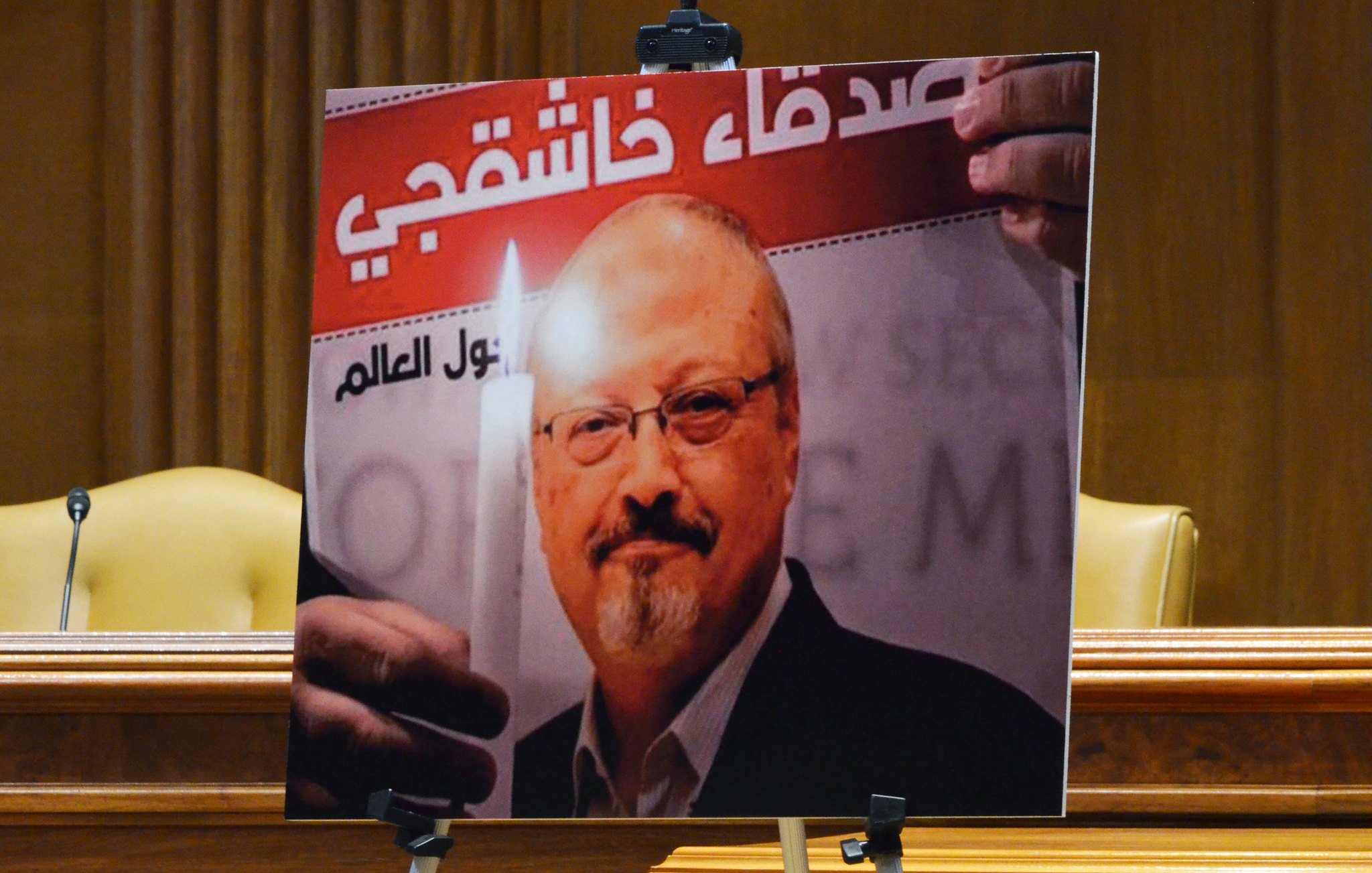 تصویر شامل پیکر مردی است که یک شمع روشن در دست دارد و با نگاهی جدی به دوربین خیره شده است. در پس‌زمینه، متنی با خط درشت وجود دارد که به موضوعی مربوط به او اشاره می‌کند. تصویر به نظر می‌رسد در یک مکان رسمی مانند یک سالن جلسات یا کنفرانس به نمایش درآمده است.