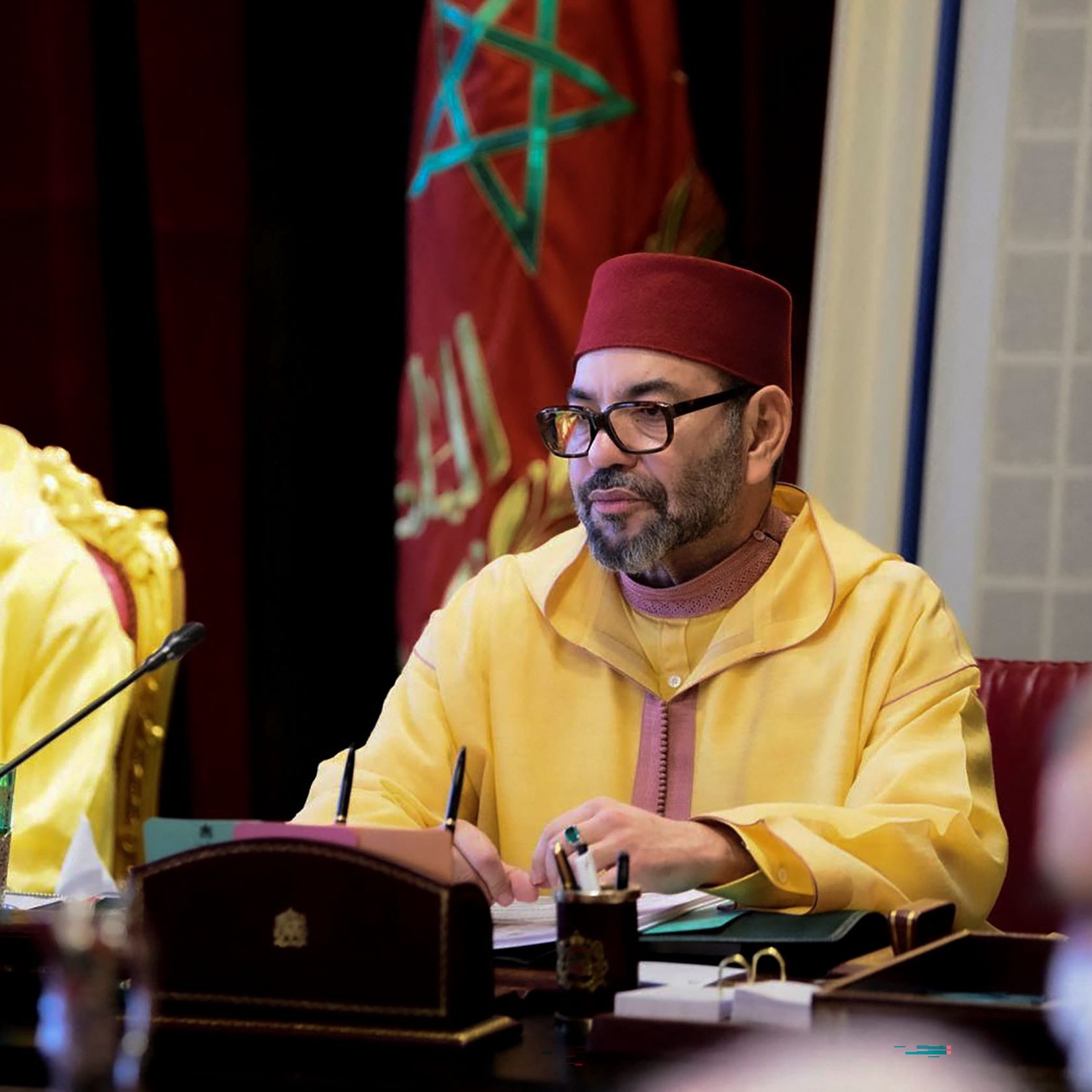 L'image montre une réunion se déroulant dans un cadre officiel. À droite, un homme vêtu d'une tenue traditionnelle marocaine de couleur jaune, portant un fez rouge, est attentif et semble concentré sur les discussions. À gauche, un jeune homme portant également un jaune et un masque facial regarde devant lui. En arrière-plan, on peut apercevoir des éléments de décoration et des drapeaux, évoquant le contexte marocain de la scène. L'ambiance générale semble sérieuse et solennelle.