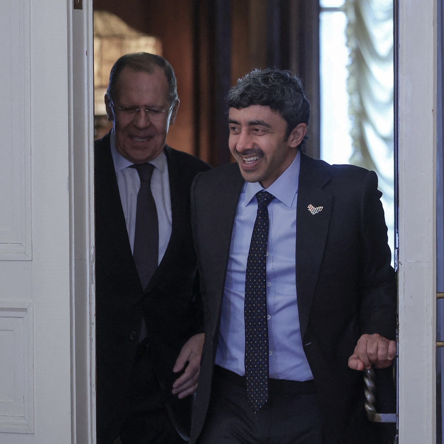 تظهر الصورة شخصين يتحدثان أثناء دخولهما من باب. الشخص في المقدمة يبدو مبتسمًا ويبدو وكأنه في حالة مزاجية جيدة، بينما الشخص الآخر خلفه يقدم تعبيرًا ودودًا. الخلفية تحتوي على تفاصيل معمارية تبرز الطابع الرسمي للمكان. كلا الشخصين يرتديان بدلات رسمية، مما يشير إلى حدث دبلوماسي أو اجتماع مهم.