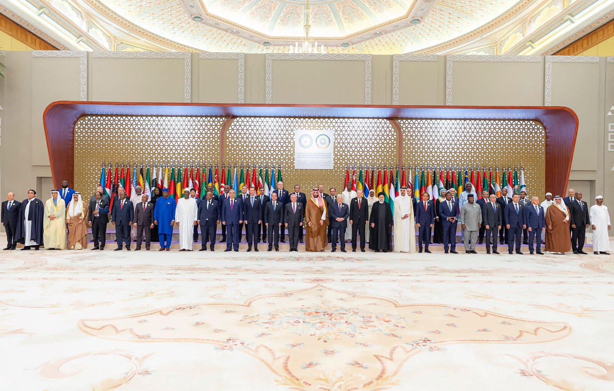 تظهر الصورة مجموعة من القادة ووزراء الخارجية المجتمعين في حدث رسمي. يقفون في صفوف مرتبة أمام خلفية مزينة بالعلم العربي، حيث تتواجد أعلام دول متعددة تشير إلى حدوث اجتماع أو قمة عربية. القادة يرتدون أزياء رسمية، بما في ذلك البذلات والعباءات، ويبدو عليهم الجدية والاهتمام بالمناسبة. الأجواء تعكس الطابع الرسمي والتاريخي لهذا اللقاء.