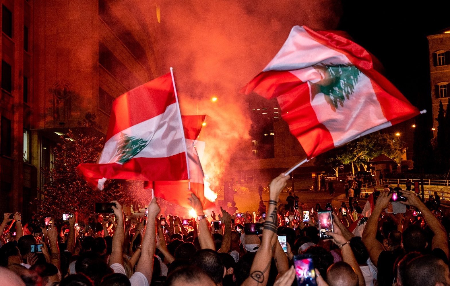 The image depicts a large gathering of people celebrating, likely in an urban setting. Many individuals are holding flags of Lebanon, which feature a red and white design with a green cedar tree. The atmosphere appears vibrant and energetic, with colorful smoke and lights adding to the scene. People are also raising their phones to capture the moment, suggesting a significant event or celebration taking place.