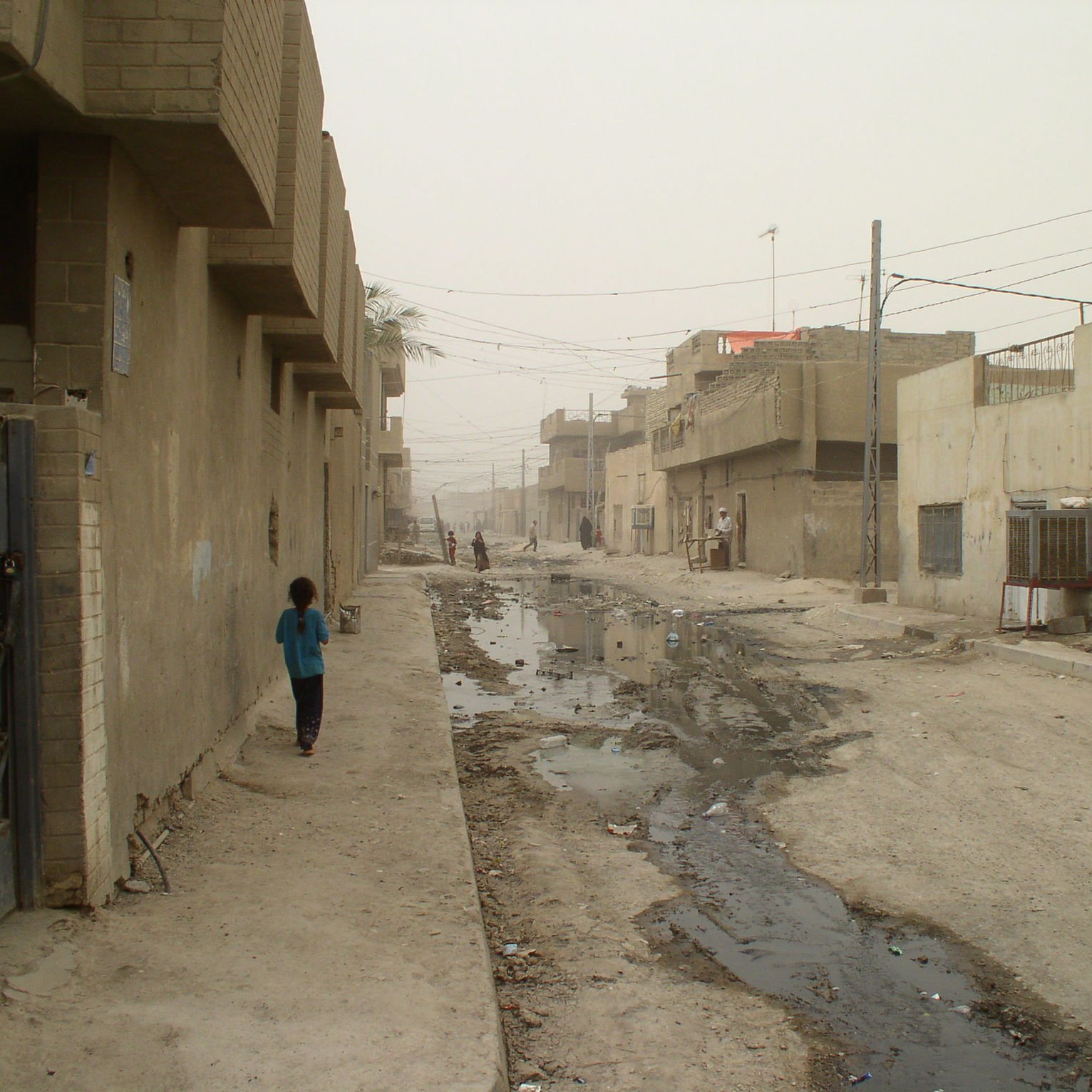الصورة تظهر شارعًا في منطقة سكنية تبدو مهملة، حيث تظهر المباني ذات اللون الرمادي والخالي من الزينة. الطريق يبدو غير معبد ويظهر فيه أجزاء من المياه والأتربة. هناك شخصان يسيران في الشارع، ورغم الغبار، فإن تفاصيل الحياة اليومية تظهر في هذه البيئة. الشارع يبدو ضيقًا وفارغًا، مما يعكس حالة من الإهمال والبساطة في التصميم العمراني.