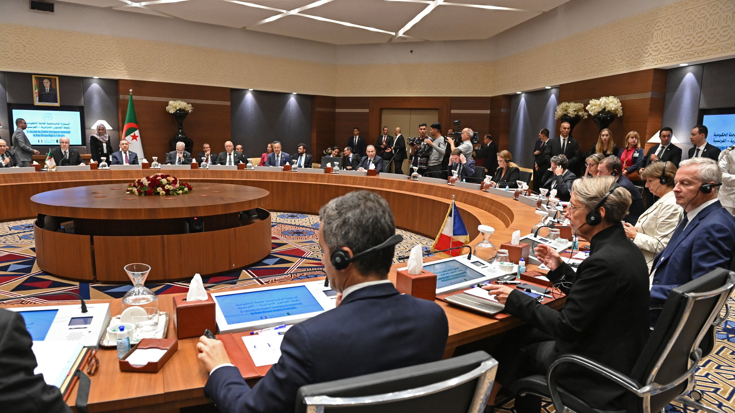 L'image montre une réunion formelle dans une salle de conférence. De nombreuses personnes sont assises autour d'une grande table ovale, probablement des représentants de différents pays ou organisations. Certains participants portent des casques d'écoute, indiquant qu'il pourrait y avoir des traductions simultanées. La salle est décorée avec des fleurs et des éléments modernes, et il y a des écrans affichant des informations pertinentes. Certaines personnes se tiennent debout en arrière-plan, observant les discussions. L'atmosphère semble professionnelle et concentrée.