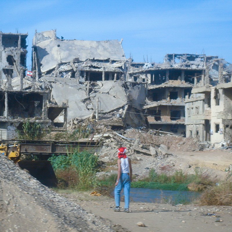 L'image montre un paysage de destruction, avec des bâtiments gravement endommagés, laissant apparaître des structures en béton et des débris éparpillés. Au premier plan, on aperçoit une personne qui semble marcher près d'une étendue d'eau, portant un chapeau rouge. L'environnement est déserté, avec une végétation qui essaie de reprendre ses droits parmi les ruines. Le ciel est bleu, mais le contraste entre la couleur du ciel et la destruction environnante crée une atmosphère de désolation.