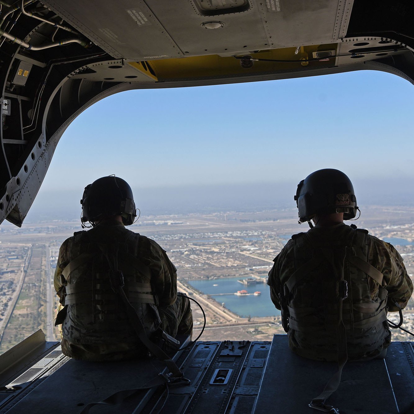 The image shows two military personnel seated inside a helicopter, looking out through the open door. They're wearing tactical gear and helmets, and the view below features a landscape with land and water. Various structures can be seen in the distance, highlighting a bustling environment beneath them. The scene conveys a sense of operation and readiness, capturing the perspective of those in flight.