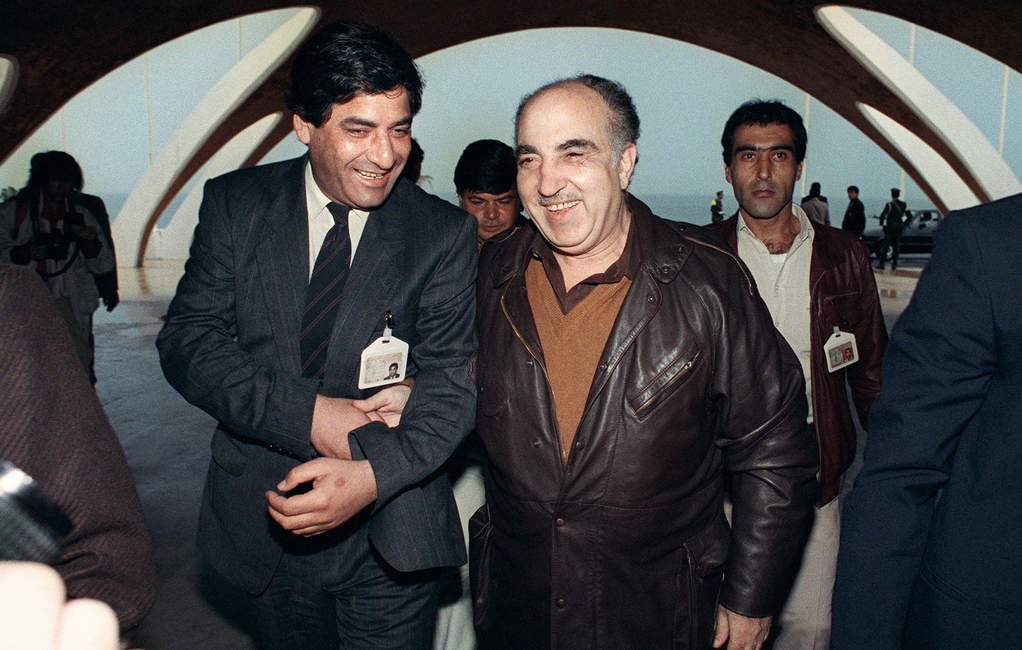 L'image montre un groupe d'hommes évoluant dans un environnement architectural moderne. Deux hommes au premier plan semblent converser de manière amicale, affichant de larges sourires. L'un est vêtu d'une veste en cuir, tandis que l'autre porte une tenue plus formelle. En arrière-plan, d'autres personnes les accompagnent, et tous semblent se déplacer sous une structure aux lignes architecturales caractéristiques. L'atmosphère générale de la photo semble être celle d'une rencontre ou d'un événement significatif.