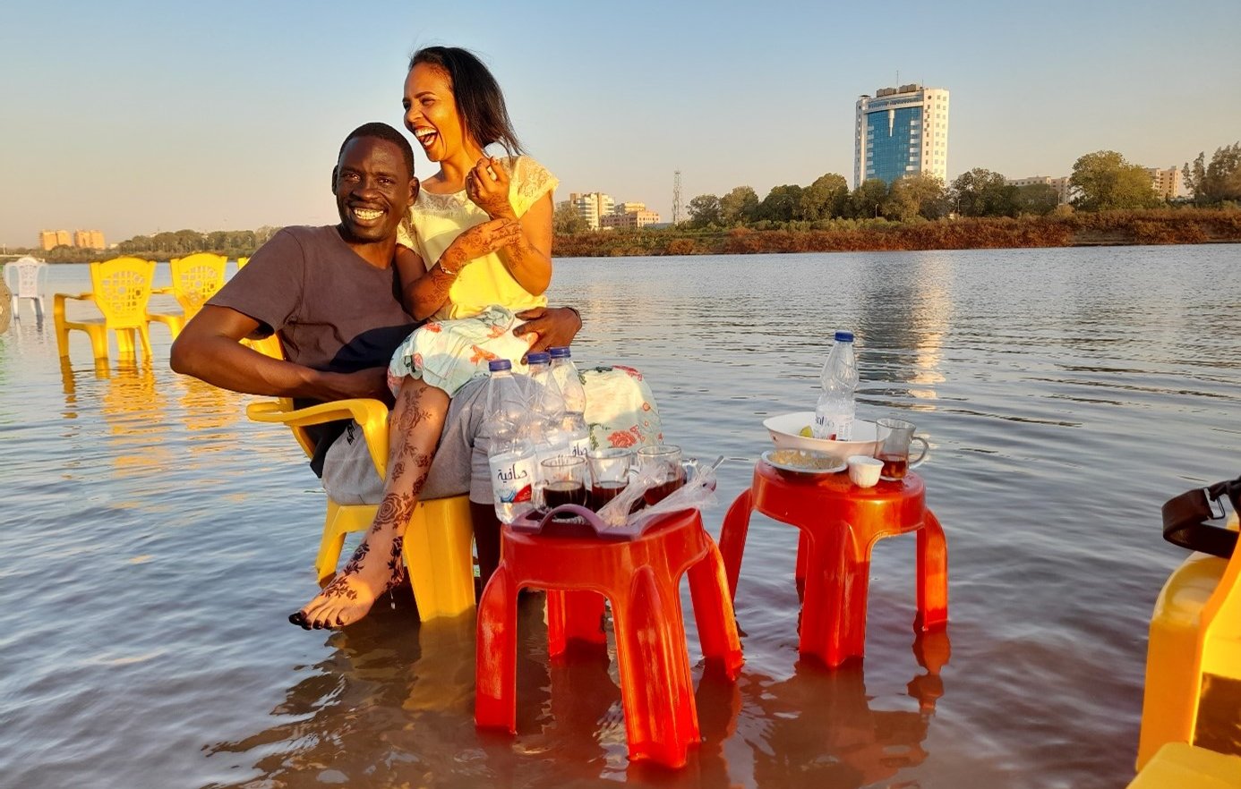 L'image montre un couple assis sur des chaises jaunes au bord d'une étendue d'eau, probablement un lac ou une rivière. Ils semblent apprécier un moment convivial ensemble, riant et partageant des instants joyeux. À côté d'eux, il y a des tables avec des bouteilles d'eau et des verres. L'arrière-plan présente un paysage urbain avec des bâtiments, et la lumière du soleil crée une atmosphère chaleureuse et accueillante.