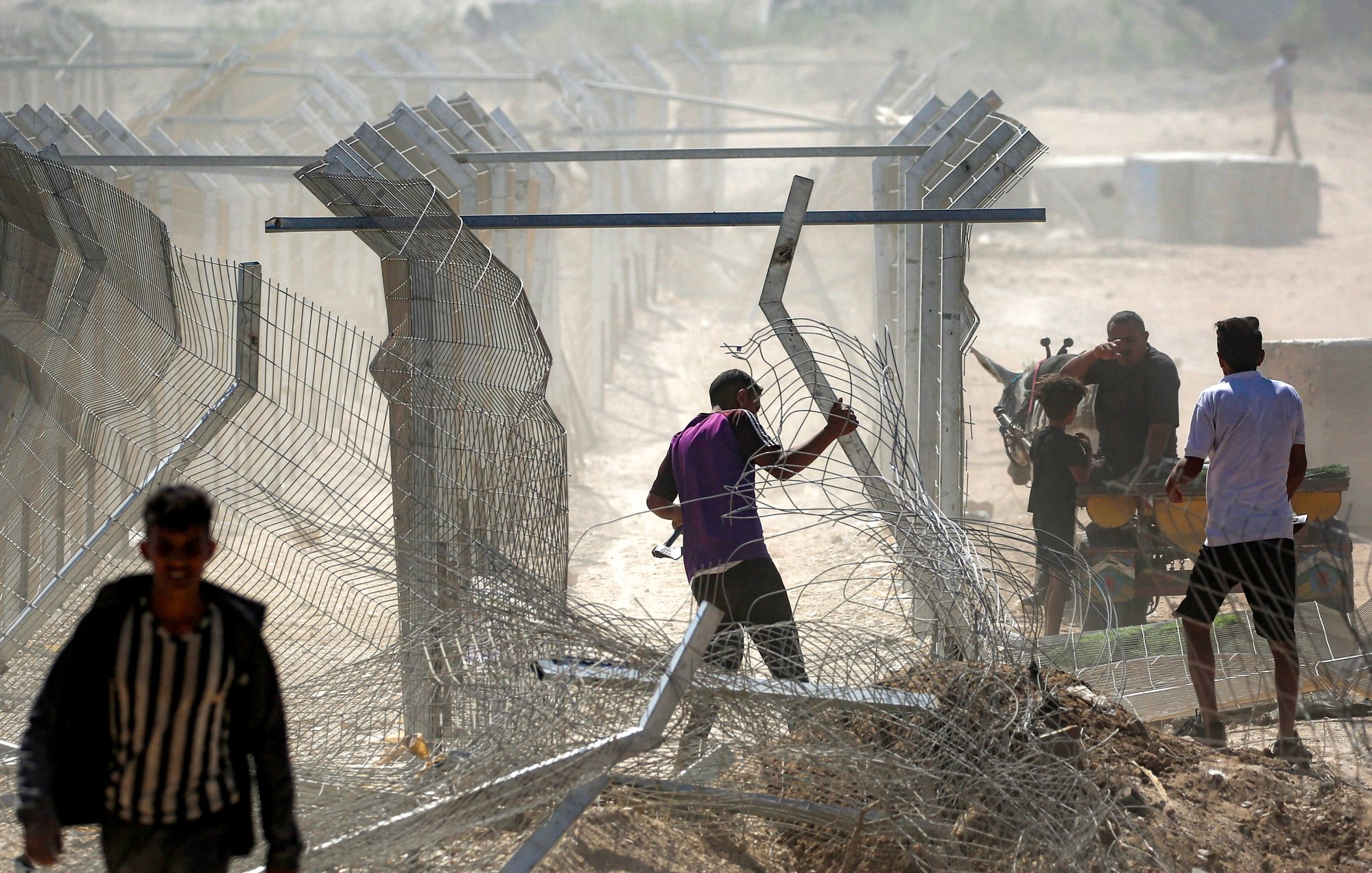 تصویر نشان‌دهنده مردانی است که در حال عبور از حصاری فلزی و برقراری یا تخریب موانع هستند. گرد و غبار در هواست.