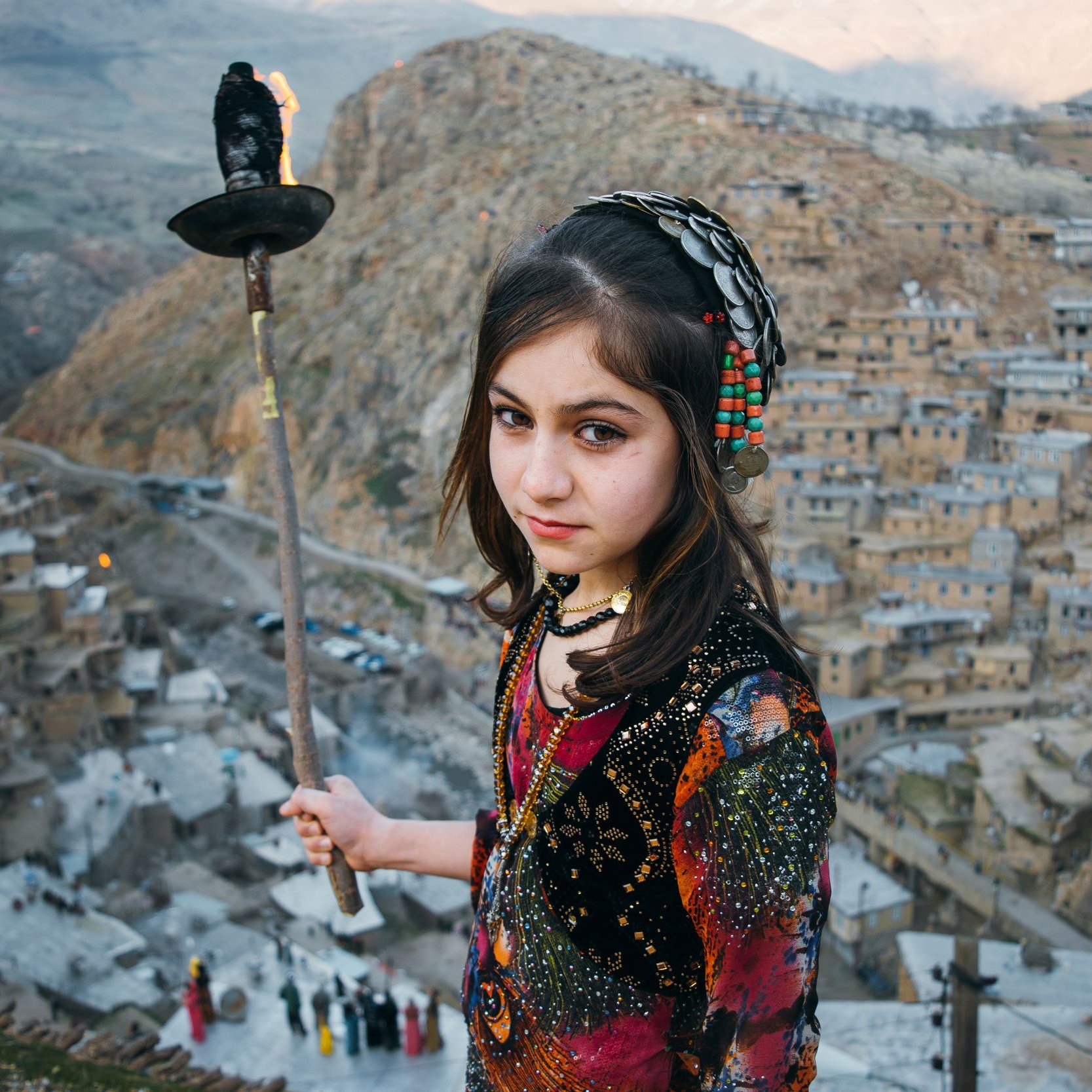 L'image montre une jeune fille tenant une torche, se tenant au sommet d'une colline surplombant un village pittoresque. Elle porte des vêtements colorés ornés de motifs traditionnels, et son regard est à la fois sérieux et perçant. En arrière-plan, on aperçoit des maisons construites à flanc de montagne, avec des paysages verdoyants s'étendant au loin. L'atmosphère de la scène est à la fois authentique et captivante, évoquant des traditions anciennes et une culture riche.