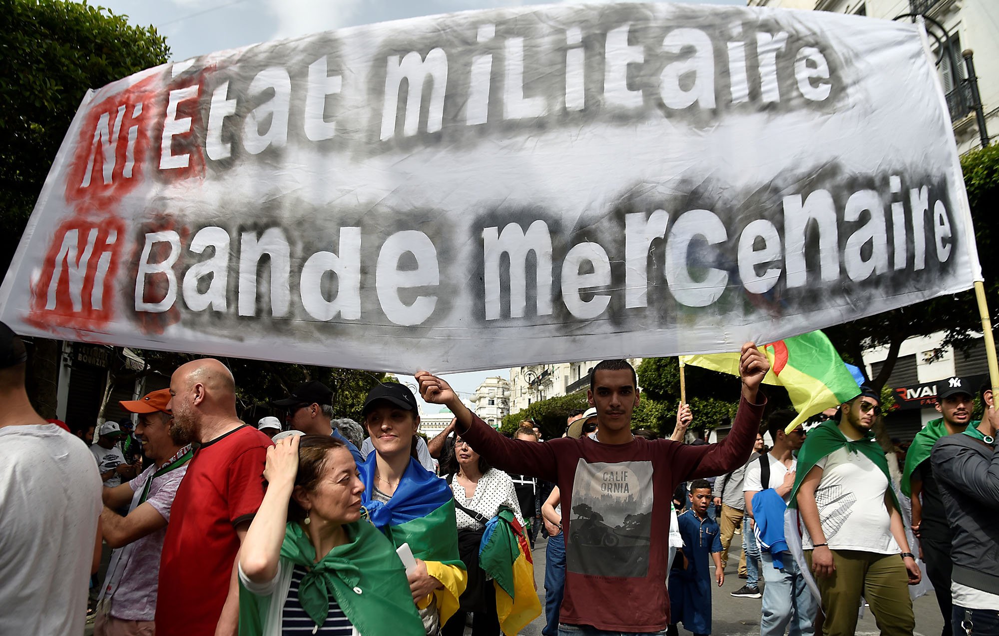 The image shows a large group of people participating in a protest or march. In the foreground, a man is holding a prominent banner that reads "Ni Etat Militaire Ni Bande Mercenaire," which translates to "Neither Military State Nor Mercenary Band." The crowd appears diverse, with individuals carrying flags and wearing various clothing styles, indicative of a collective demonstration. The background features urban scenery, suggesting the protest is taking place in a city.