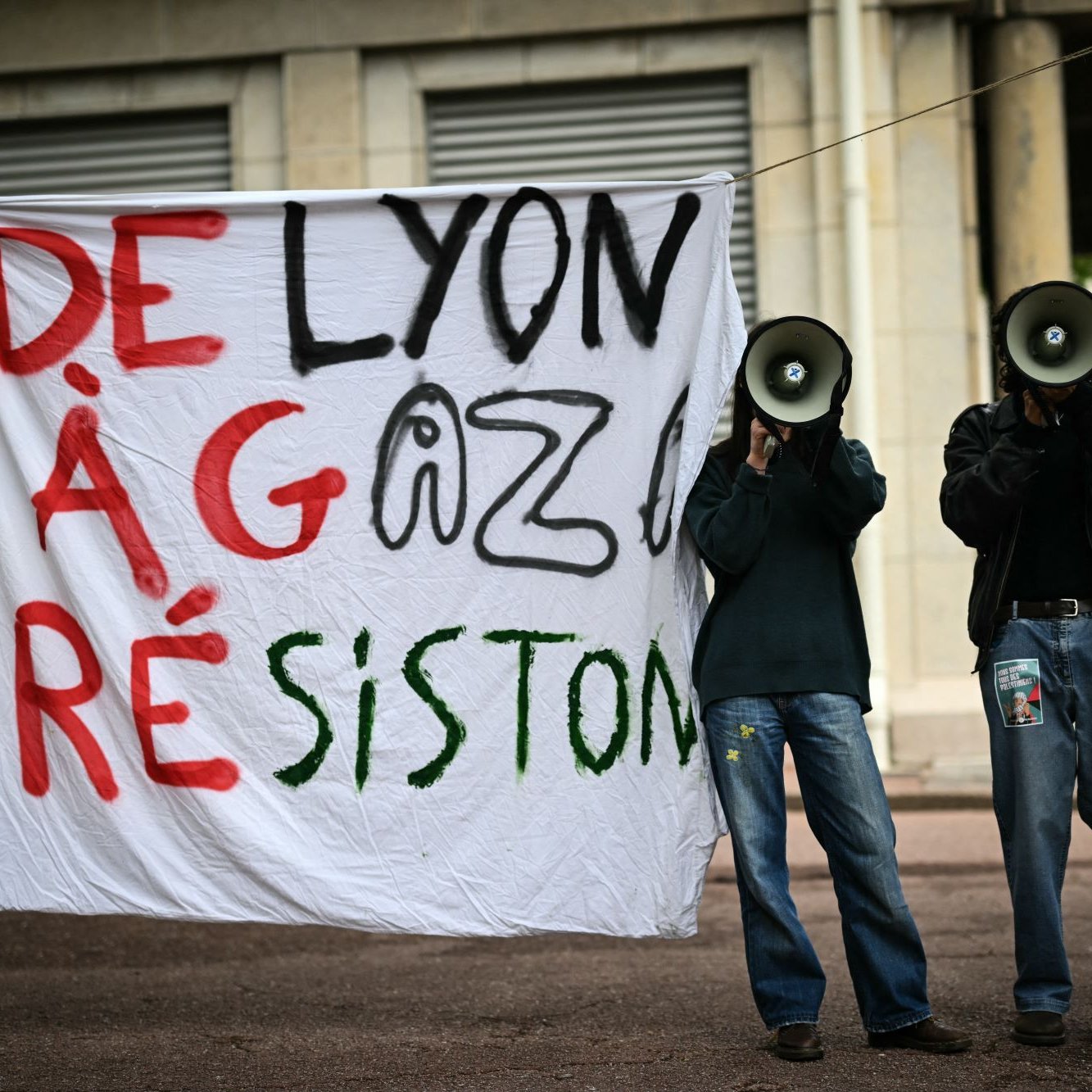 L'image montre une manifestation avec une grande pancarte accrochée qui dit "DE LYON À GAZA RÉSISTONS". Deux personnes se tiennent derrière la pancarte, chacune tenant un mégaphone. L'environnement semble urbain, et l'atmosphère suggère un engagement pour une cause.