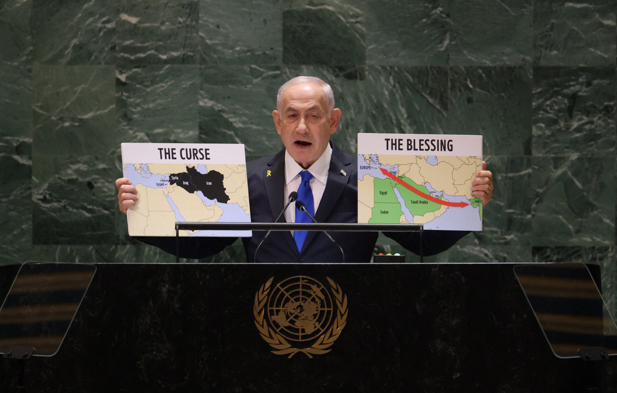 L'image montre un homme debout derrière un pupitre, s'exprimant lors d'un discours. Il tient deux panneaux, l'un portant l'inscription "THE CURSE" (la malédiction) et l'autre "THE BLESSING" (la bénédiction). Sur ces panneaux, il y a des cartes représentant des régions du Moyen-Orient, probablement pour illustrer un point sur des questions géopolitiques. Le décor est formel, semblant indiquer une réunion internationale, comme celle des Nations Unies. L'homme porte une cravate et une veste, se tenant de manière convaincante.