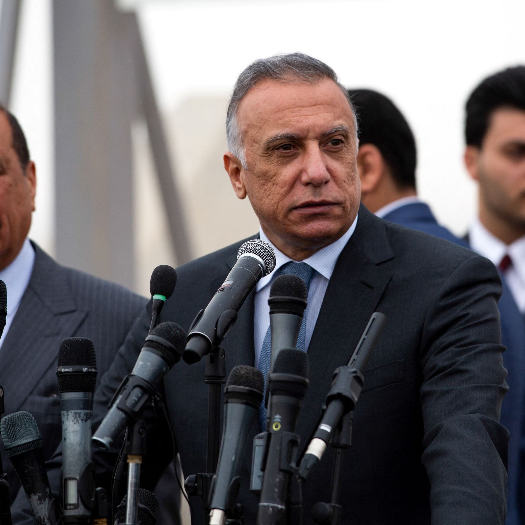 تظهر الصورة مجموعة من الرجال أثناء مؤتمر صحفي. في المقدمة يوجد رجل يرتدي بدلة رسمية، ويبدو جادًا وهو يتحدث أو يستعد للإدلاء ببيان. خلفه، يوجد رجل آخر يرتدي بدلة أيضًا، يبدو أنه يستمع أو يشارك في الحوار. هناك عدد من الميكروفونات أمامهم، مما يوحي بأنهم في موقف يتطلب إلقاء الخطابات أو التصريحات الإعلامية. الجو العام يبدو رسميًا، وقد تكون هناك بعض الضبابية في الخلفية.