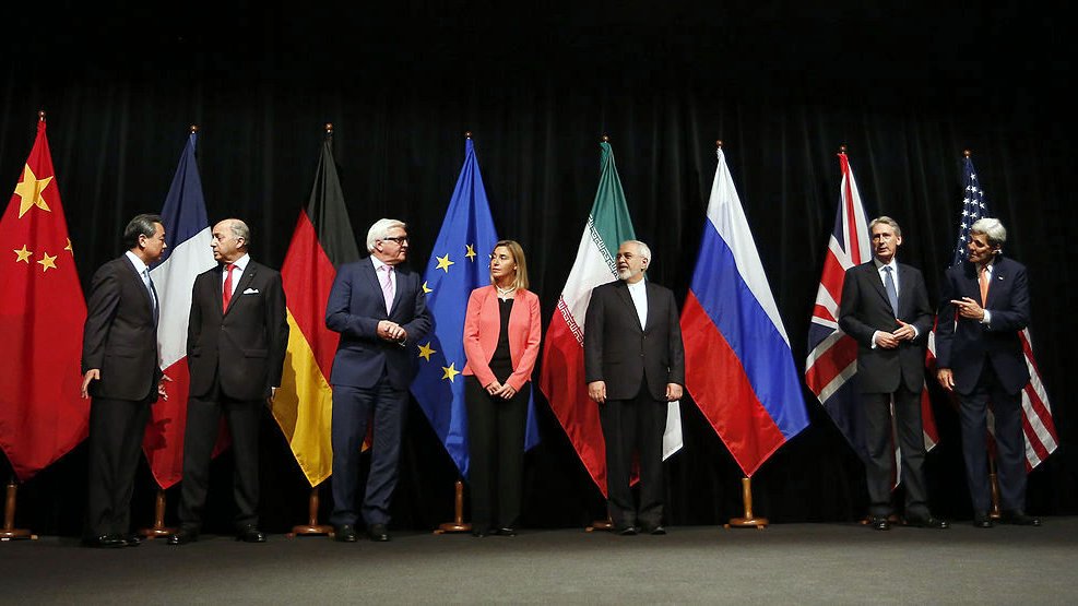 تظهر الصورة مجموعة من القادة أو الممثلين السياسيين واقفين أمام مجموعة من الأعلام لدول مختلفة، تشمل الصين، ألمانيا، الاتحاد الأوروبي، إيران، روسيا، وبريطانيا. يبدو أن الصورة قد تم التقاطها خلال حدث رسمي، حيث يتحدث بعضهم بينما ينظر الآخرون بشكل جاد. الأعلام مرتبة بشكل منظم، مما يعكس سياق سياسي دولي أو تفاوضي.
