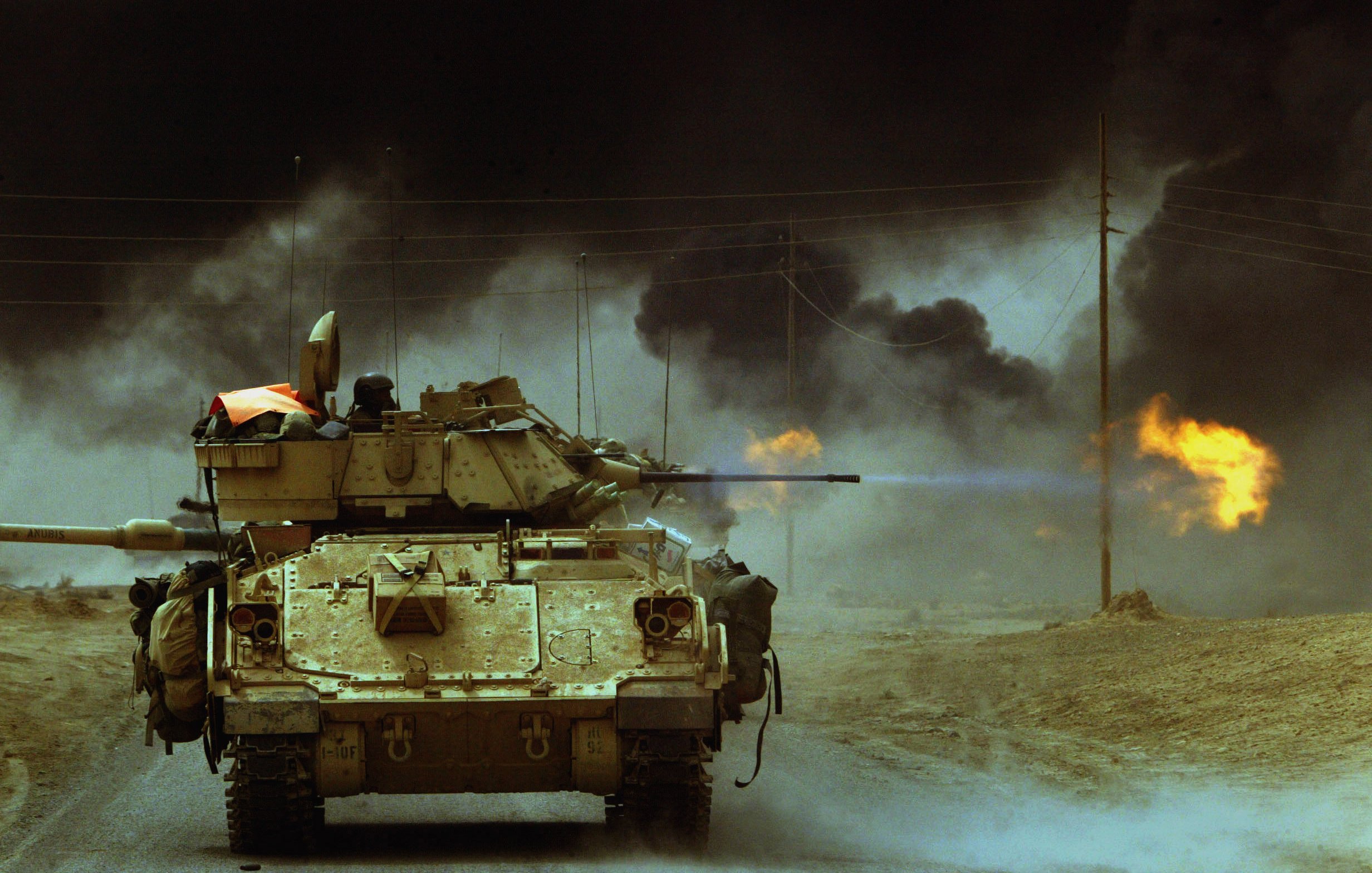 L'image montre un char de guerre sur une route, se dirigeant vers l'avant. En arrière-plan, on aperçoit des nuages de fumée noire qui s'échappent de flammes, suggérant un environnement de conflit ou d'escalade militaire. La scène est sombre et dramatique, évoquant une ambiance de tension et de danger. Le char est équipé d'un canon, et l'ensemble de l'image dépeint une situation de guerre.