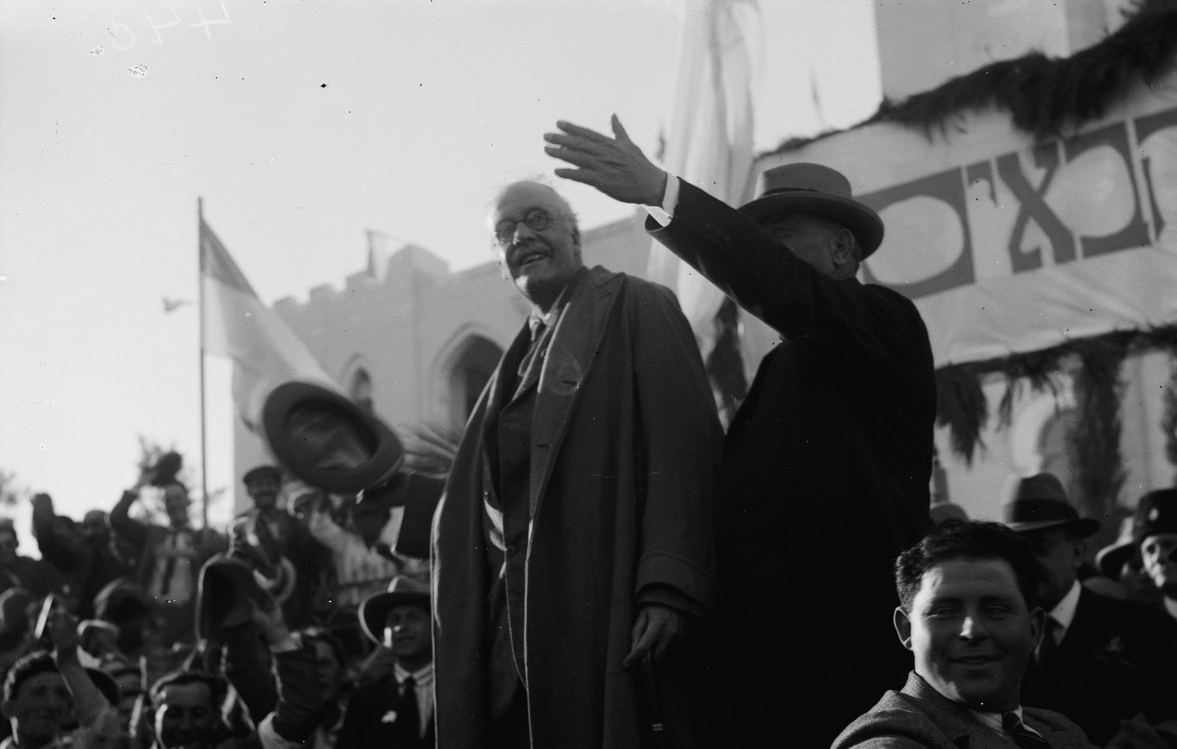L'image montre des hommes en costume debout sur une estrade, saluant une foule rassemblée. L'un des hommes, portant un manteau, semble s'adresser au public, tandis que l'autre, en chapeau, lève la main. En arrière-plan, on aperçoit des drapeaux et des décorations, indiquant un événement officiel ou une célébration. La foule est composée de personnes applaudissant et prenant part à l'événement. L'atmosphère semble festive et solennelle.