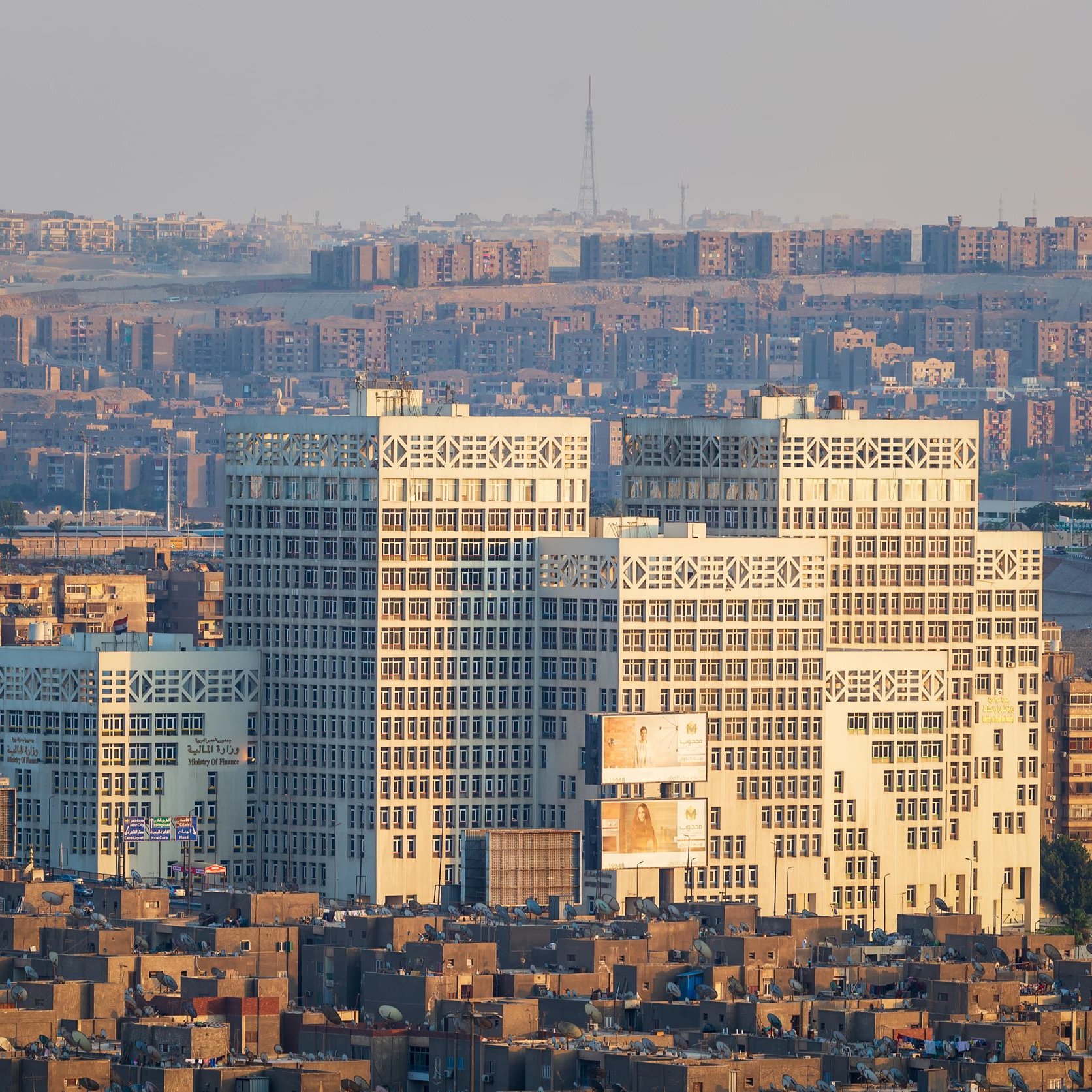 تظهر في الصورة مدينة تتميز بكثافة المباني والشقق السكنية. هناك مجموعة من المباني الحديثة ذات التصميم الفريد في المقدمة، حيث ينعكس عليها ضوء الشمس مما يبرز تفاصيلها. خلفها، تظهر منطقة سكنية كثيفة البناء، مع مشهد لجوانب مختلفة من المدينة. يبدو أن الجو هادئ، مع سماء مضيئة وقليل من الضباب.