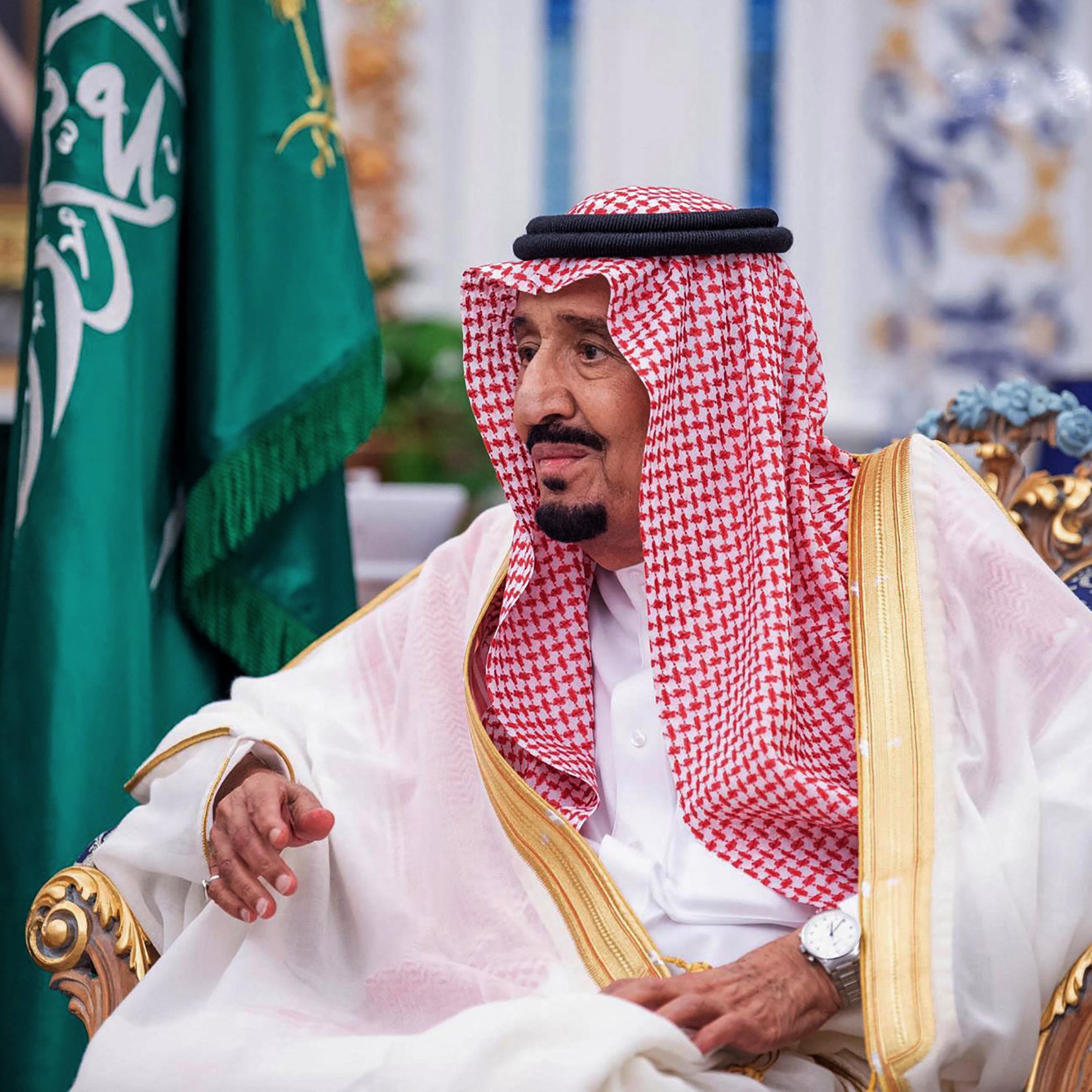 The image shows a man seated in a lavish decor room, wearing traditional attire that includes a white thobe and a red and white checked keffiyeh held in place by an agal. He has a thoughtful expression and is gesturing with his hand. Behind him, there is a green flag, likely representing Saudi Arabia, and opulent furnishings in the background.