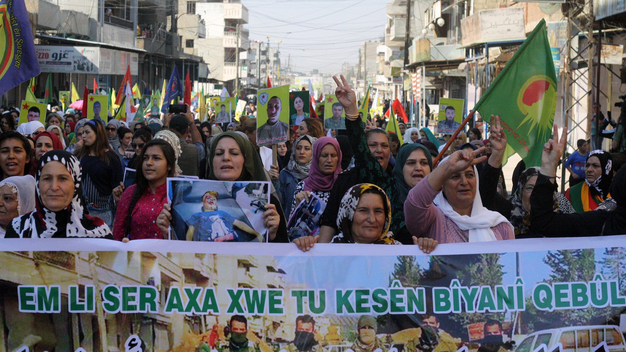 L'image montre une grande foule de personnes, principalement des femmes, participant à une manifestation. Elles portent des drapeaux colorés et affichent des pancartes avec des slogans. Les manifestants semblent exprimer des revendications et un soutien, probablement liées à des questions politiques ou sociales. L'ambiance est dynamique et déterminée, avec des signes de solidarité parmi les participants. Les rues environnantes sont visibles, ainsi que des bâtiments en arrière-plan.