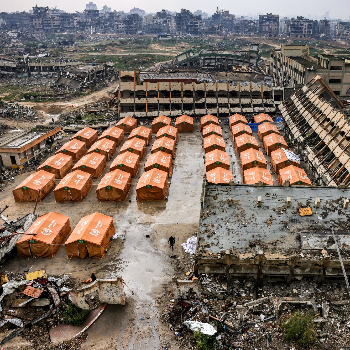 این تصویر یک منطقه ویران را نشان می‌دهد که شامل تعدادی چادر نارنجی رنگ است. در پس‌زمینه، ساختمان‌های تخریب شده و زمین‌های خالی دیده می‌شوند. به نظر می‌رسد این مکان به عنوان محل اسکان موقت استفاده می‌شود. فضای کلی تصویر حس خرابی و ضرورت کمک را منتقل می‌کند.
