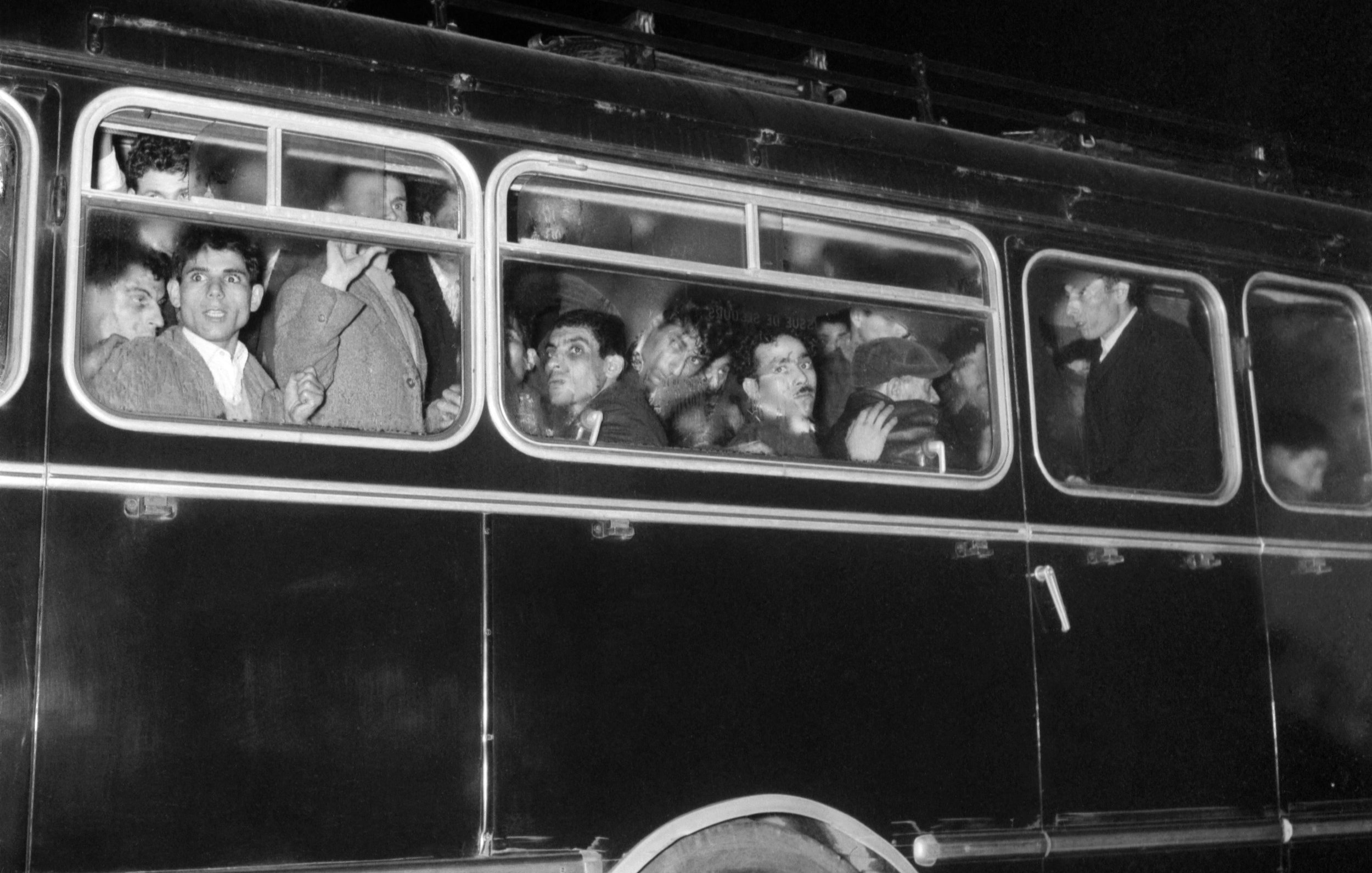 The image depicts a crowded bus at night. Several people are visible through the windows, some looking out while others appear to be engaged in conversation. The bus is dark, possibly painted black, and has a classic design with multiple windows. The expressions of the individuals range from curious to contemplative, highlighting a moment of shared experience among the passengers. The scene captures a sense of movement and typical urban life.