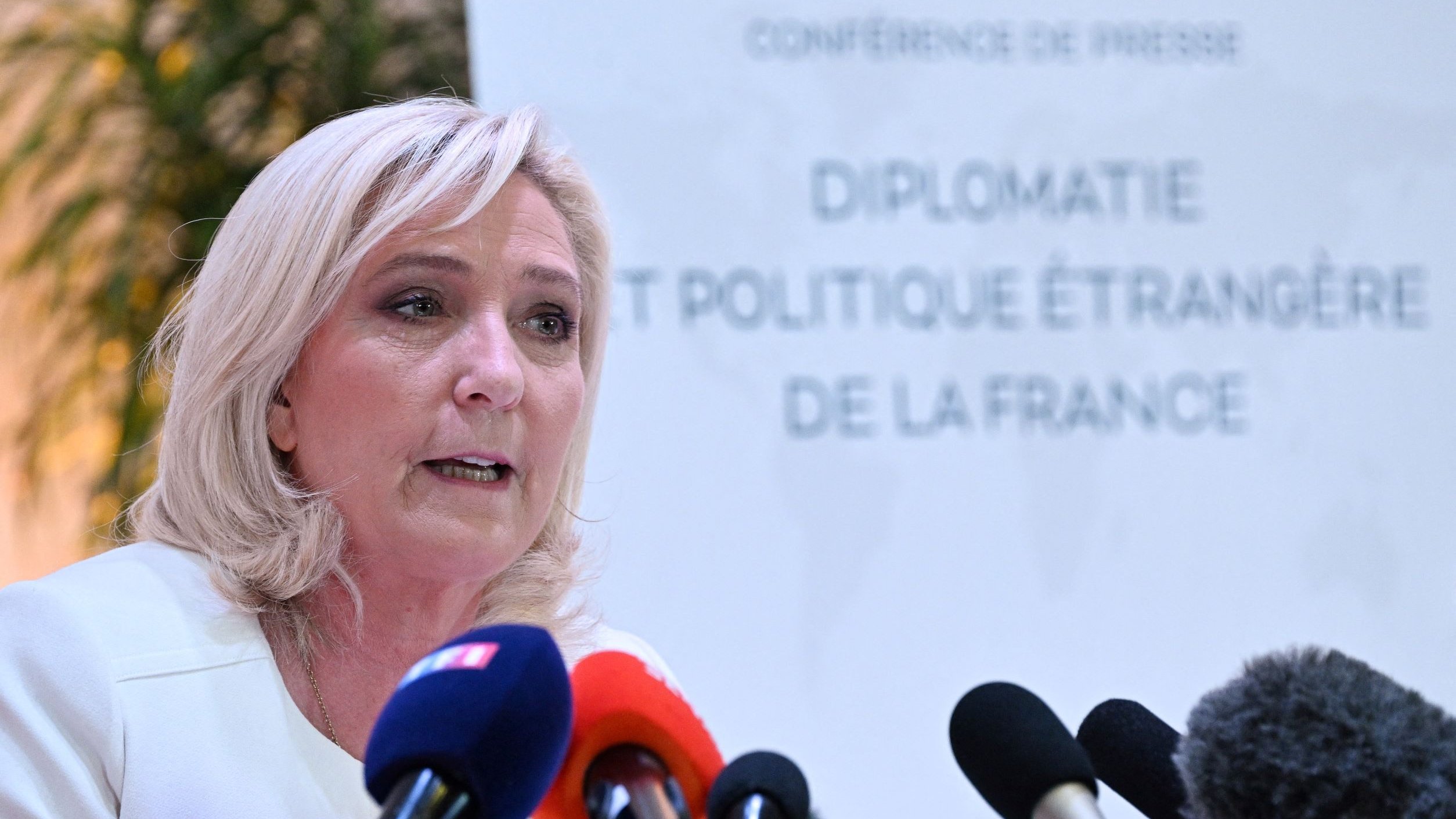 L'image montre une femme qui s'exprime lors d'une conférence de presse. Elle a des cheveux blonds et porte une veste claire. Au fond, on peut voir un panneau qui indique "CONFERENCE DE PRESSE" ainsi que le titre "DIPLOMATIE ET POLITIQUE ÉTRANGÈRE DE LA FRANCE". Plusieurs microphones sont disposés devant elle, suggérant qu'elle s'adresse aux médias. L'ambiance semble formelle et professionnelle.